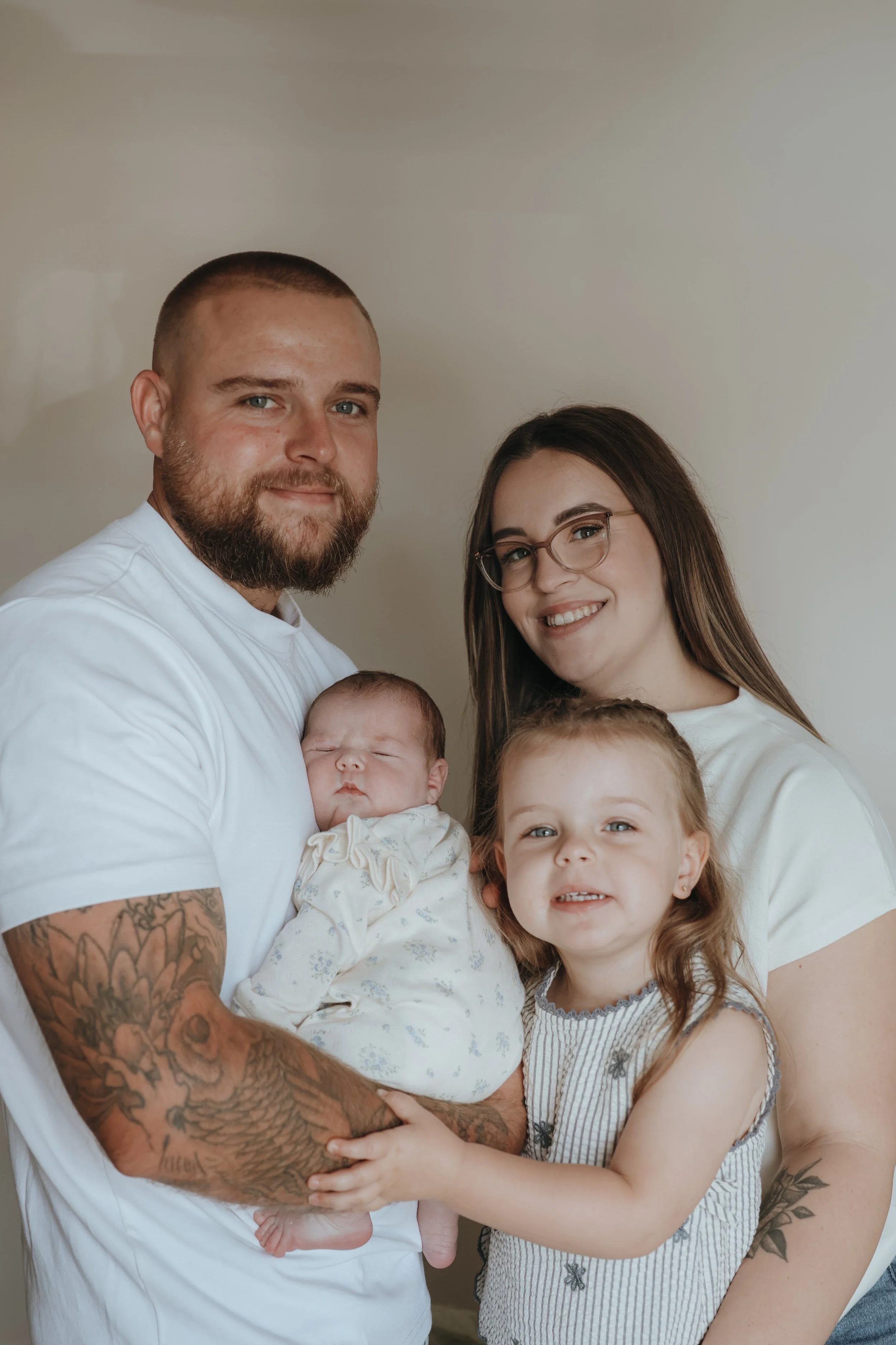 A man with tattoos, a woman with glasses, and three children, including one baby, are posing together indoors with a neutral wall background.
