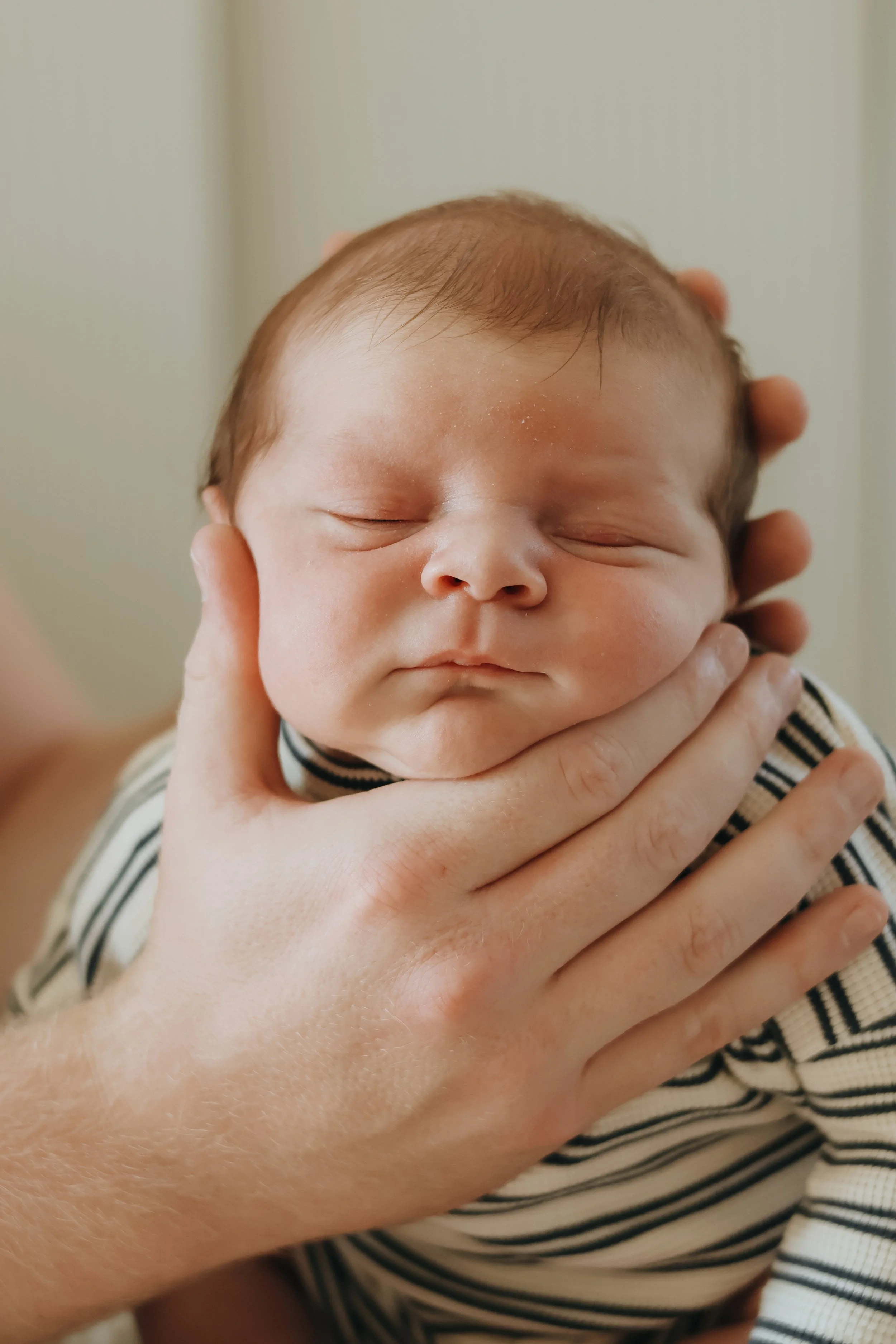 Close-up of a newborn baby with closed eyes, being gently supported by an adult's hand, wearing a striped outfit.
