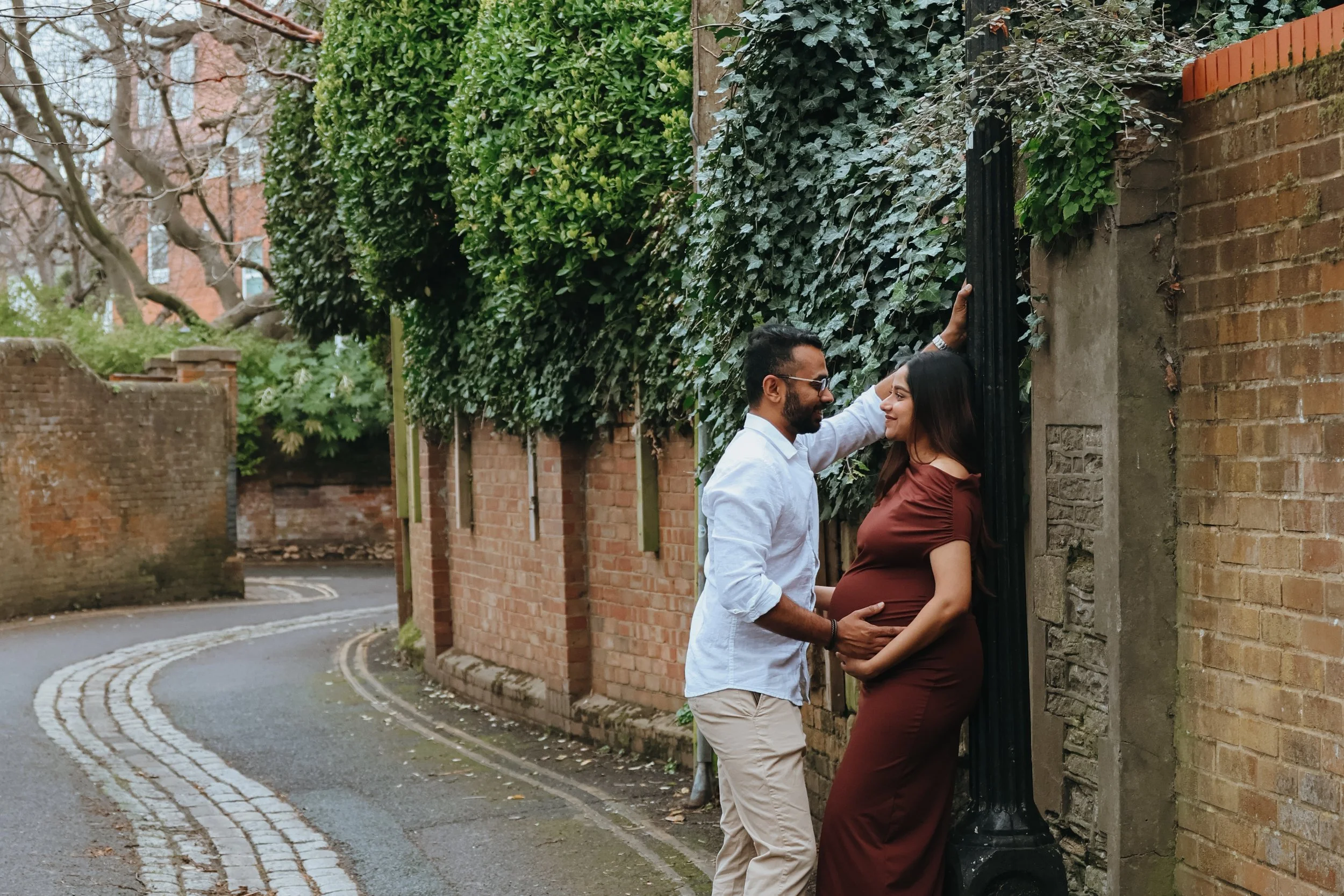 A couple stands close together on a narrow, winding street, with the man gently touching the wall behind the woman, and both are smiling. The woman is pregnant, wearing a brown dress, and the man is dressed in a white shirt and beige pants. Ivy cover