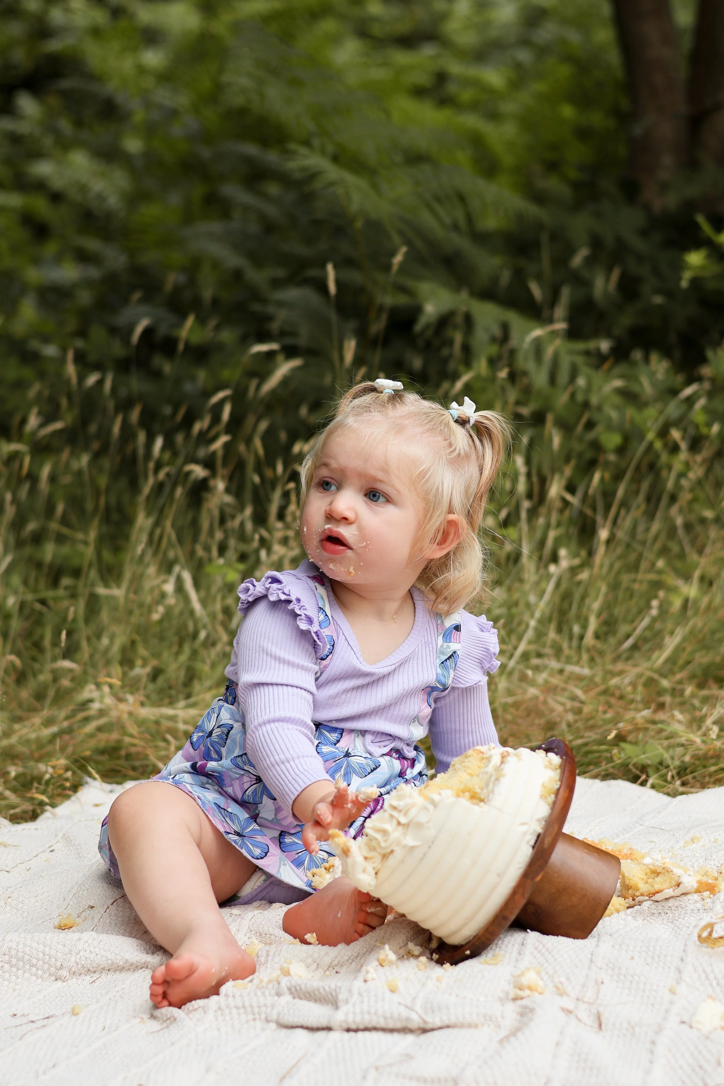 A young girl sitting on a blanket outdoors with cake in front of her, surrounded by grass and trees. cake smash, first birthday photography, southampton 