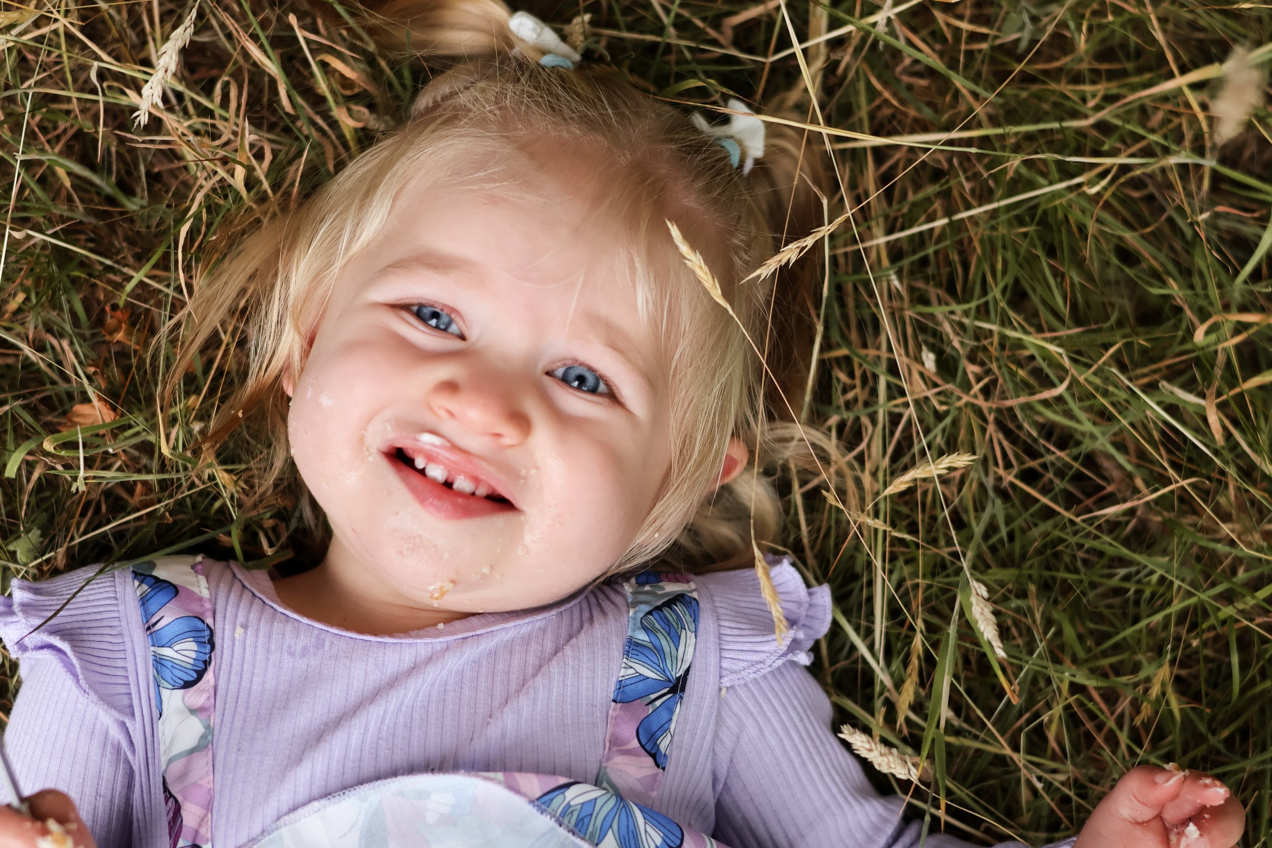A smiling young girl with blue eyes and blonde hair lying on her back in grass, with cake and food remnants on her face and clothes. cake smash, first birthday photography, Hampshire 