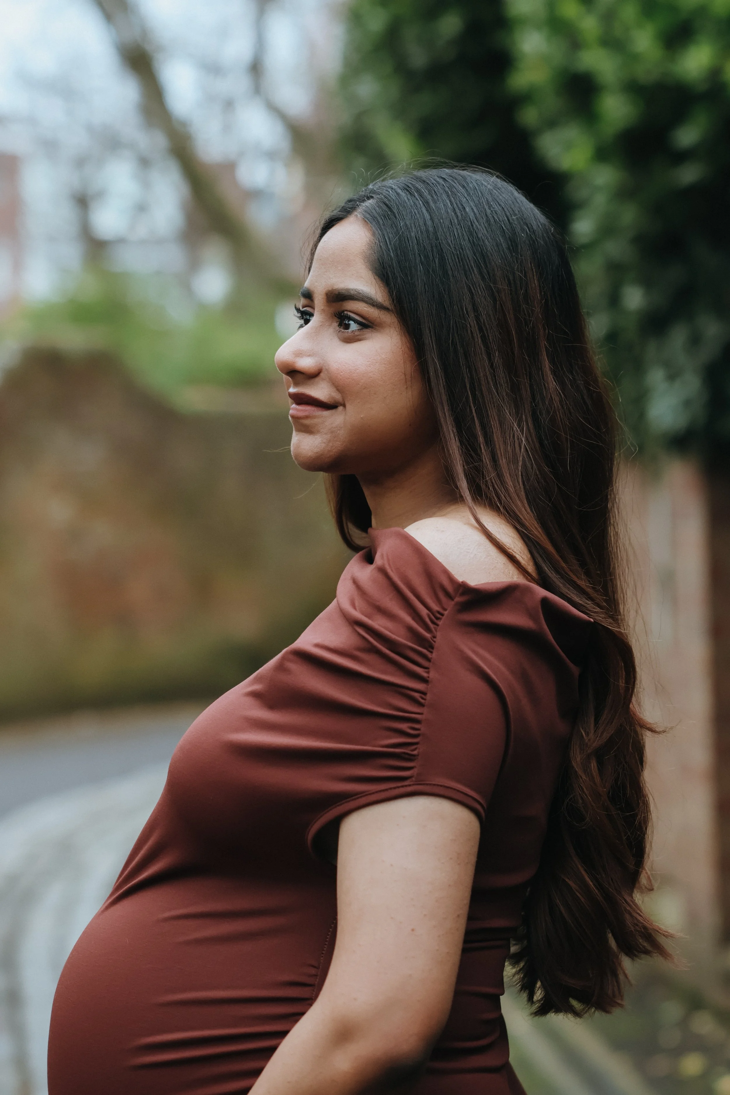 A pregnant woman with long dark hair standing outdoors in front of a blurred background of trees and a brick wall, wearing a fitted brown dress with puffed sleeves. Maternity session, maternity photography, Portsmouth 