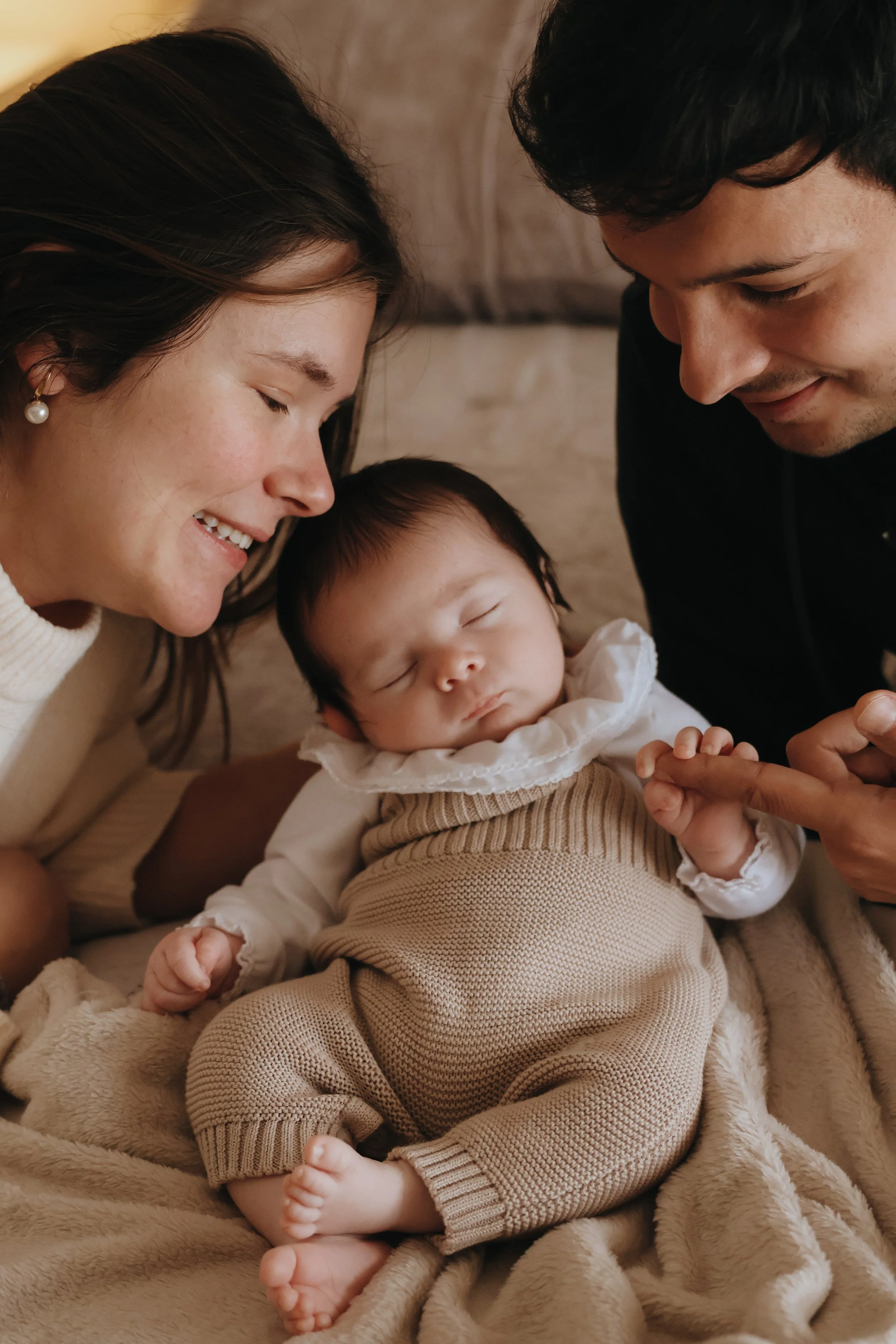 A couple with their sleeping baby,  the mother smiling and leaning towards the baby, and the father looking down at the baby with a gentle expression, all lying on a soft beige blanket.
