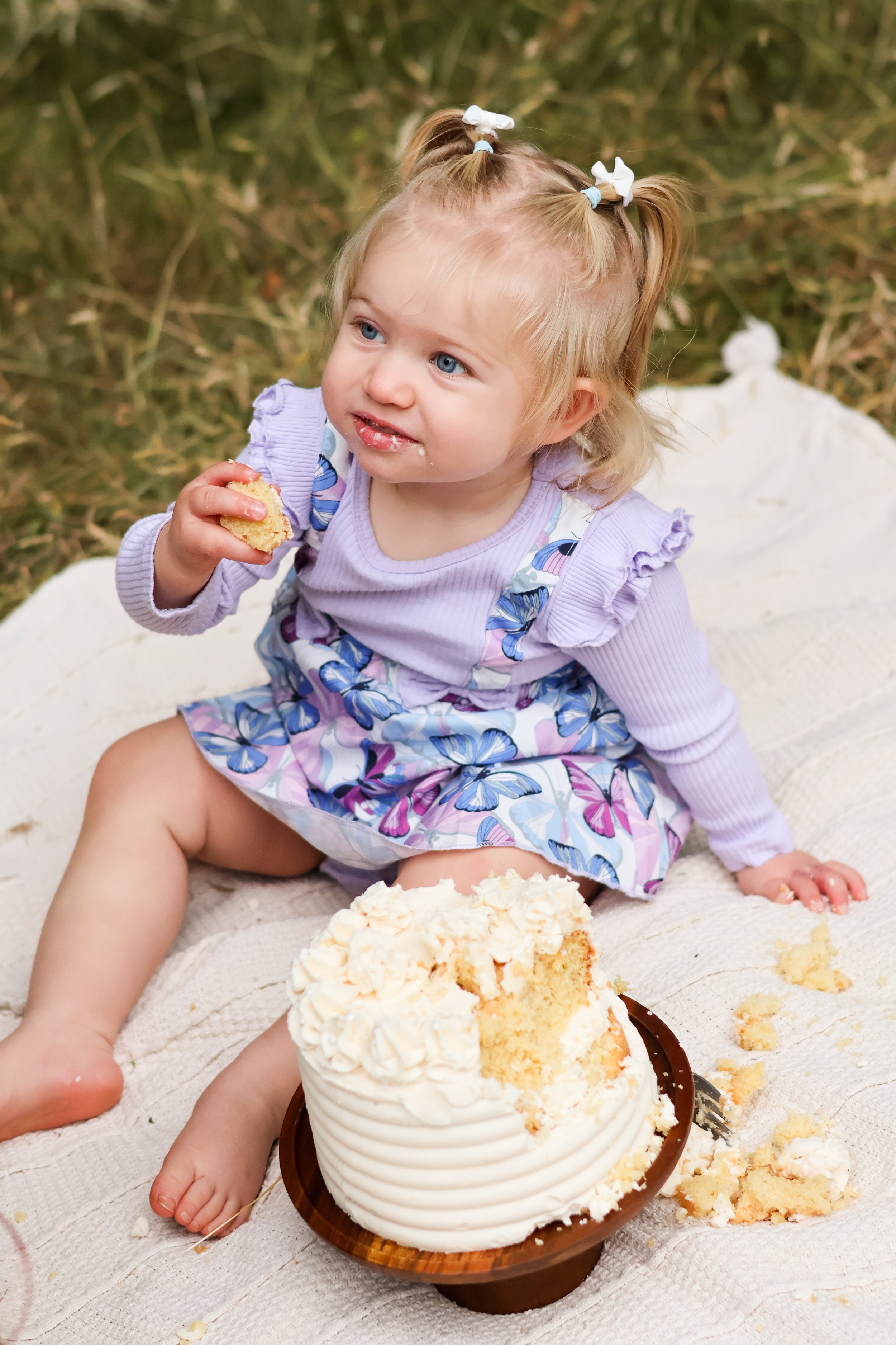 A young girl with blonde hair in pigtails and blue eyes, wearing a butterfly-patterned dress and lavender sweater, sitting on a white blanket outdoors, eating cake.