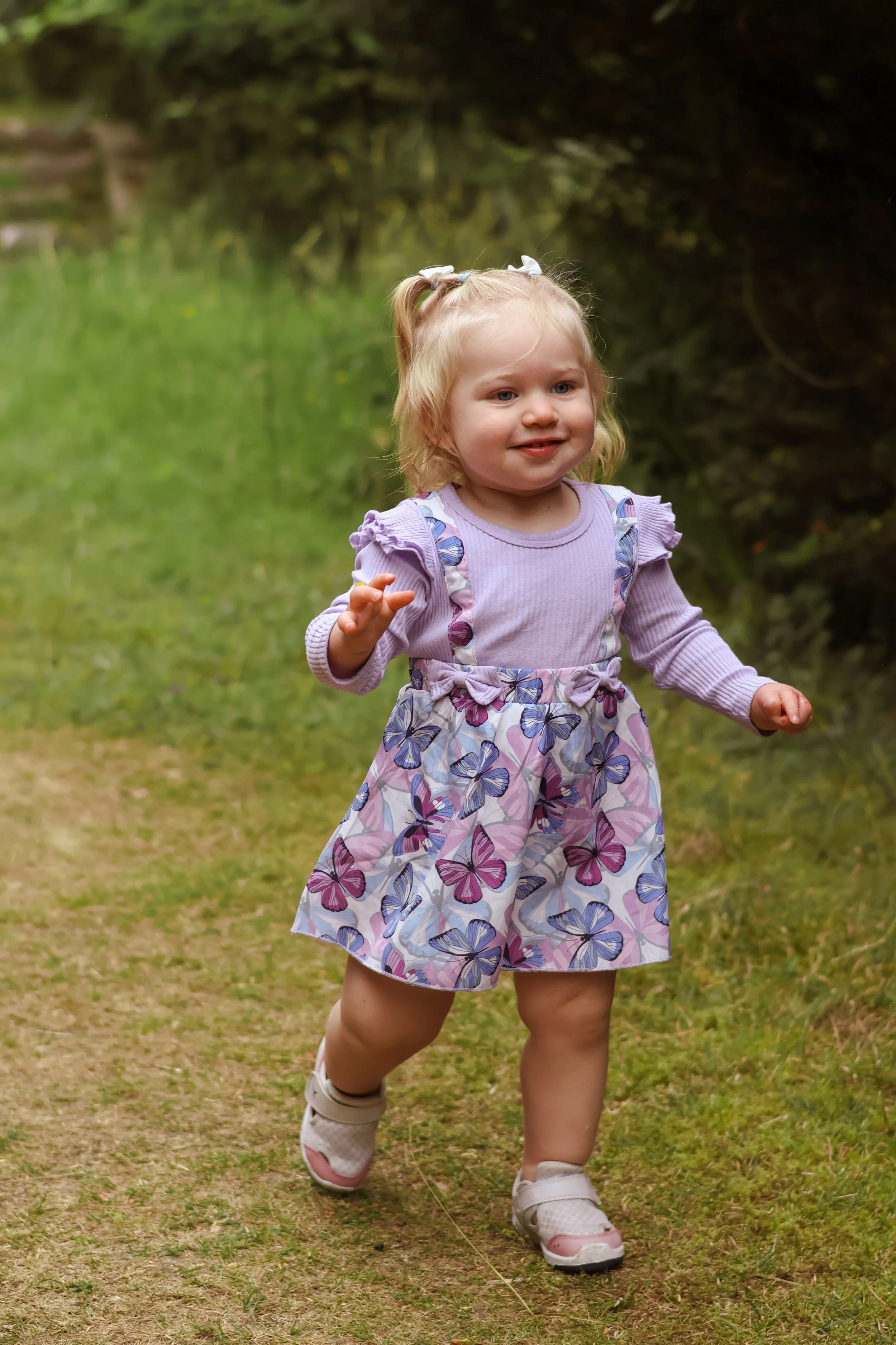 A young girl with blonde hair in pigtails, wearing a purple long-sleeve shirt and a butterfly-patterned dress, running outdoors on grass.