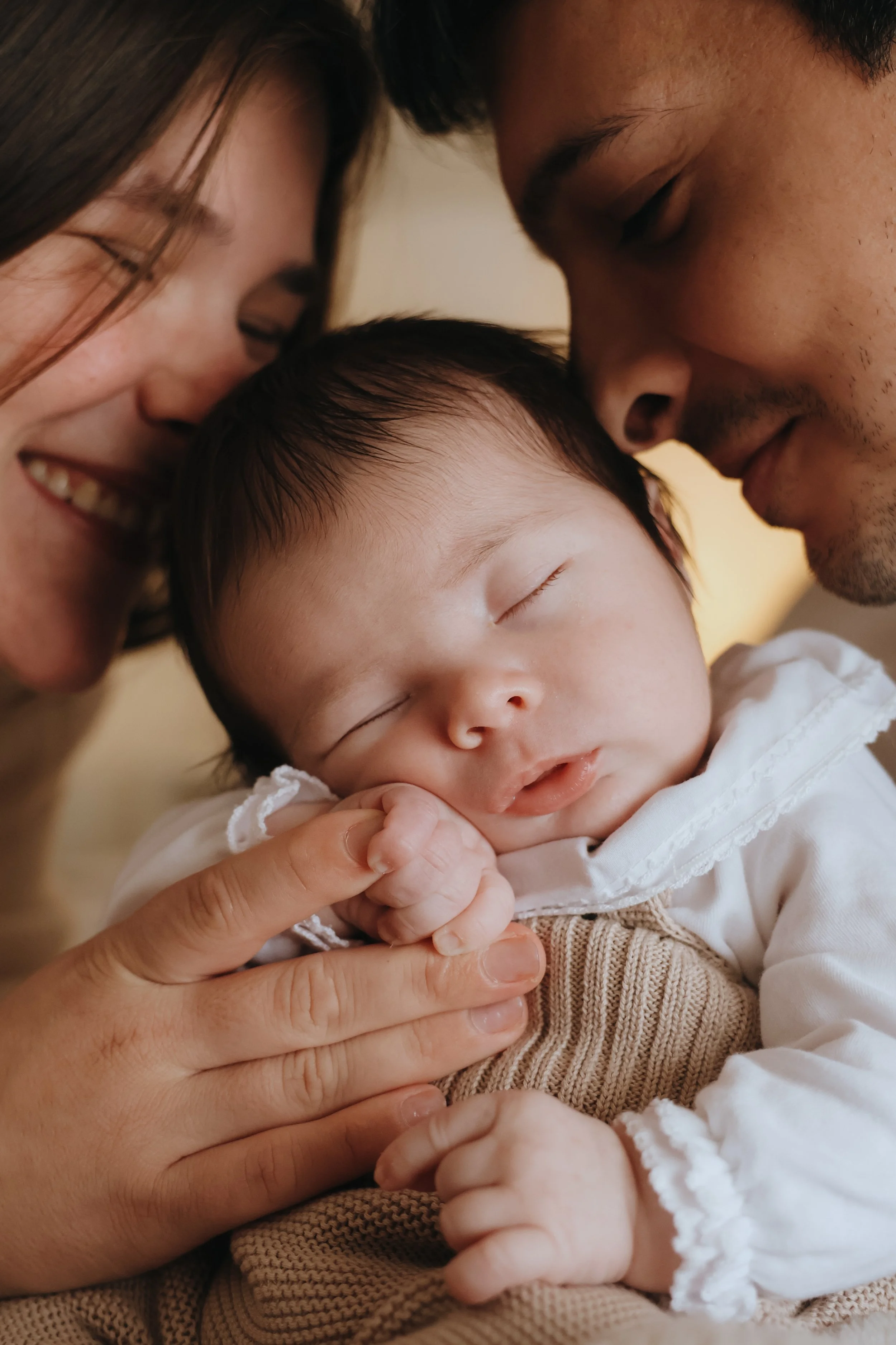 A family of three, a woman, a man, and a sleeping baby, close together with their foreheads touching, showing affection.