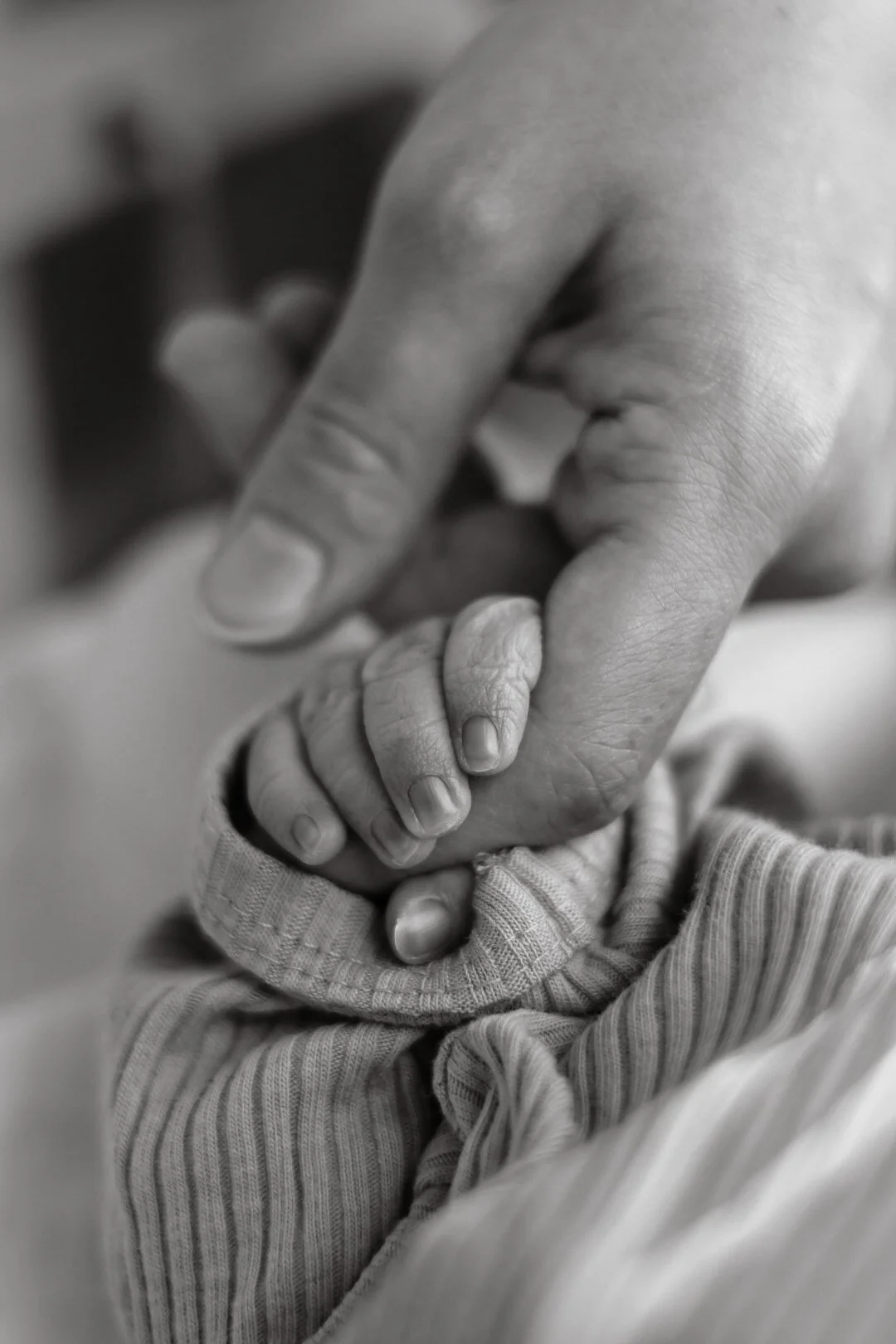Close-up of an adult hand gently holding a baby's hand, both showing detailed textures, in black and white.