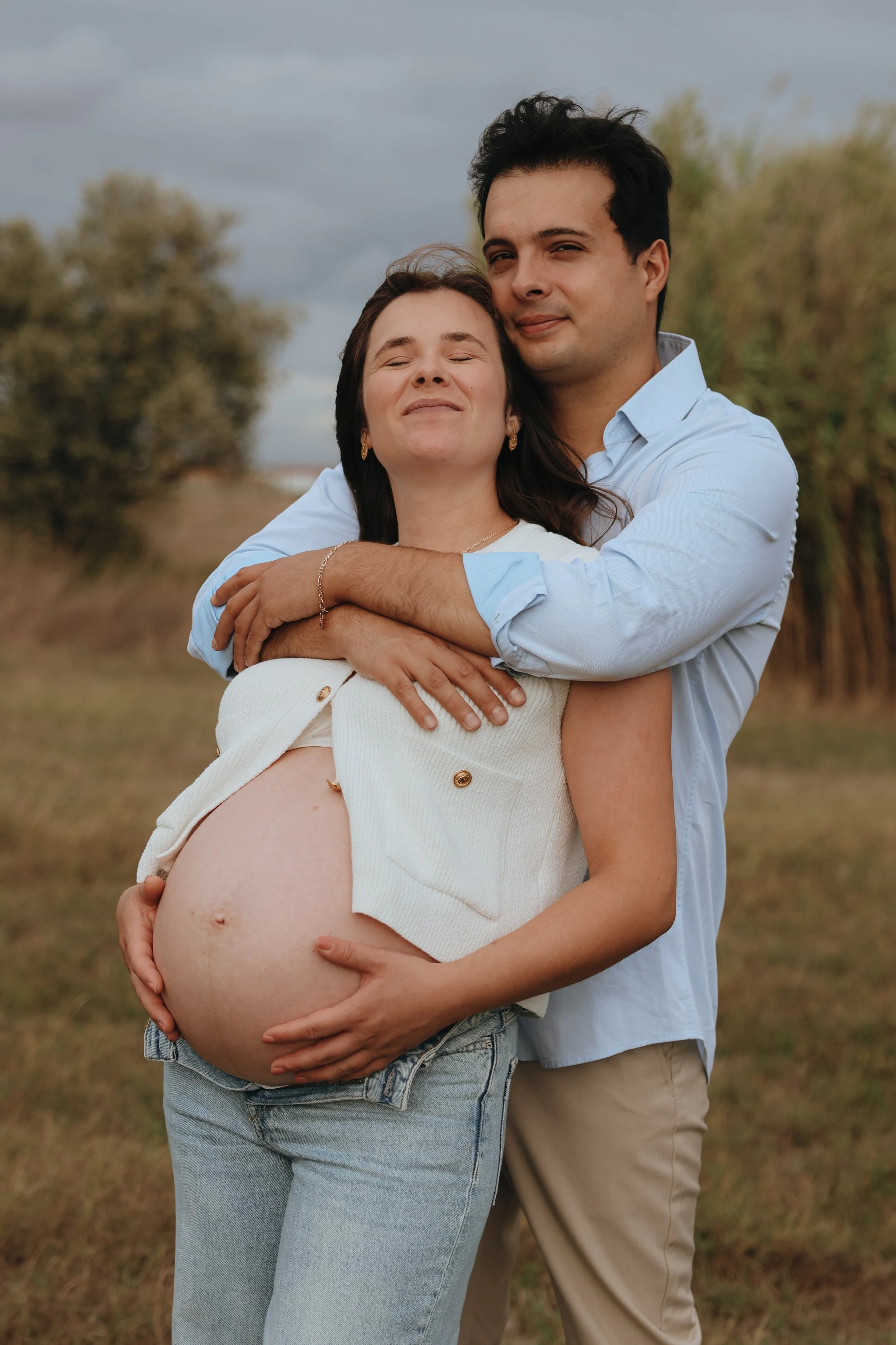 A pregnant woman and a man are standing outdoors, with the man hugging the woman from behind. The woman has a content expression and is holding her pregnant belly. The scene is set in a grassy area with trees in the background and a cloudy sky overhe