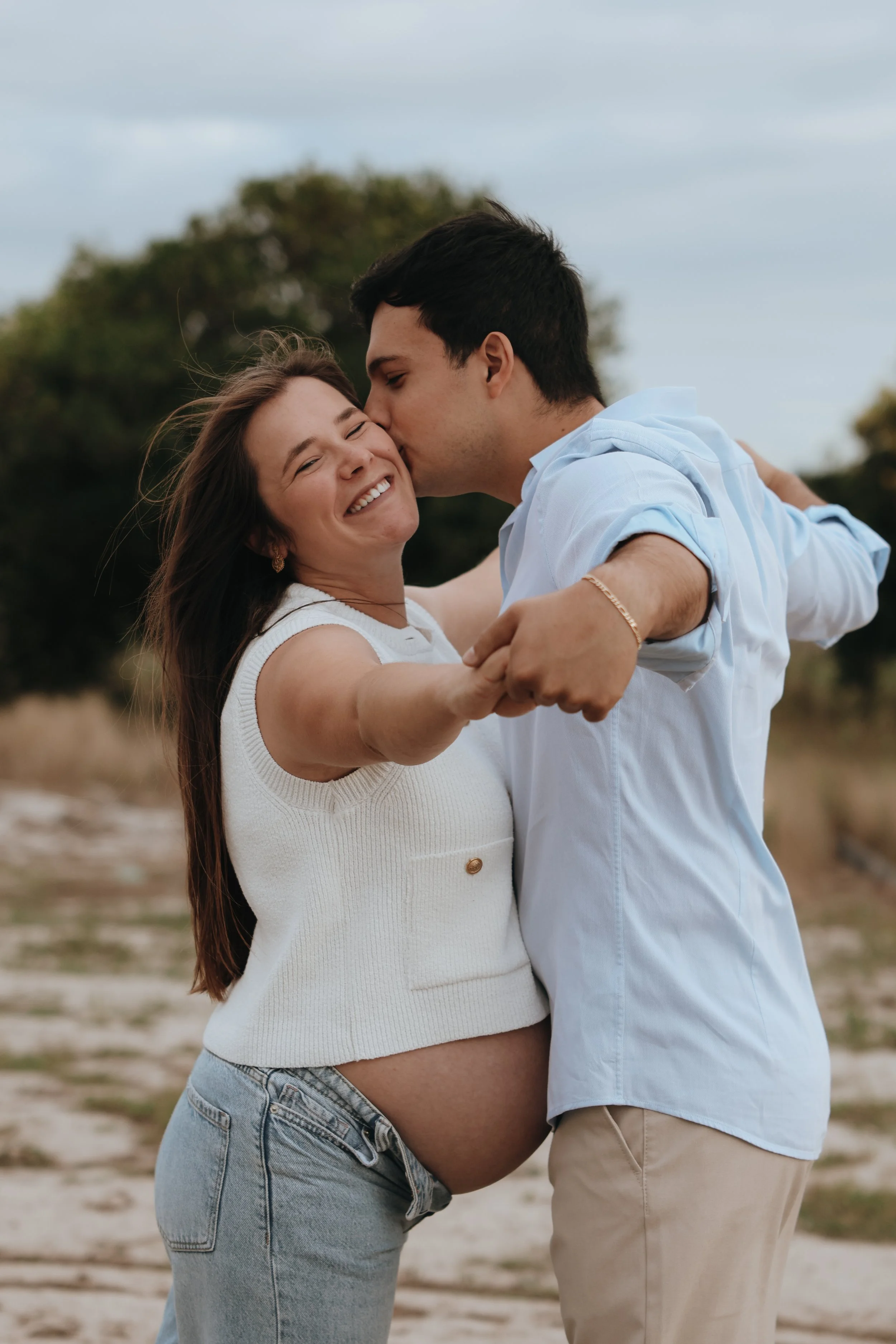A pregnant woman and a man are embracing outdoors, with the man kissing the woman on the forehead. The woman is smiling and appears happy, standing on a sandy path with trees in the background. Maternity session outdoor Hampshire 
