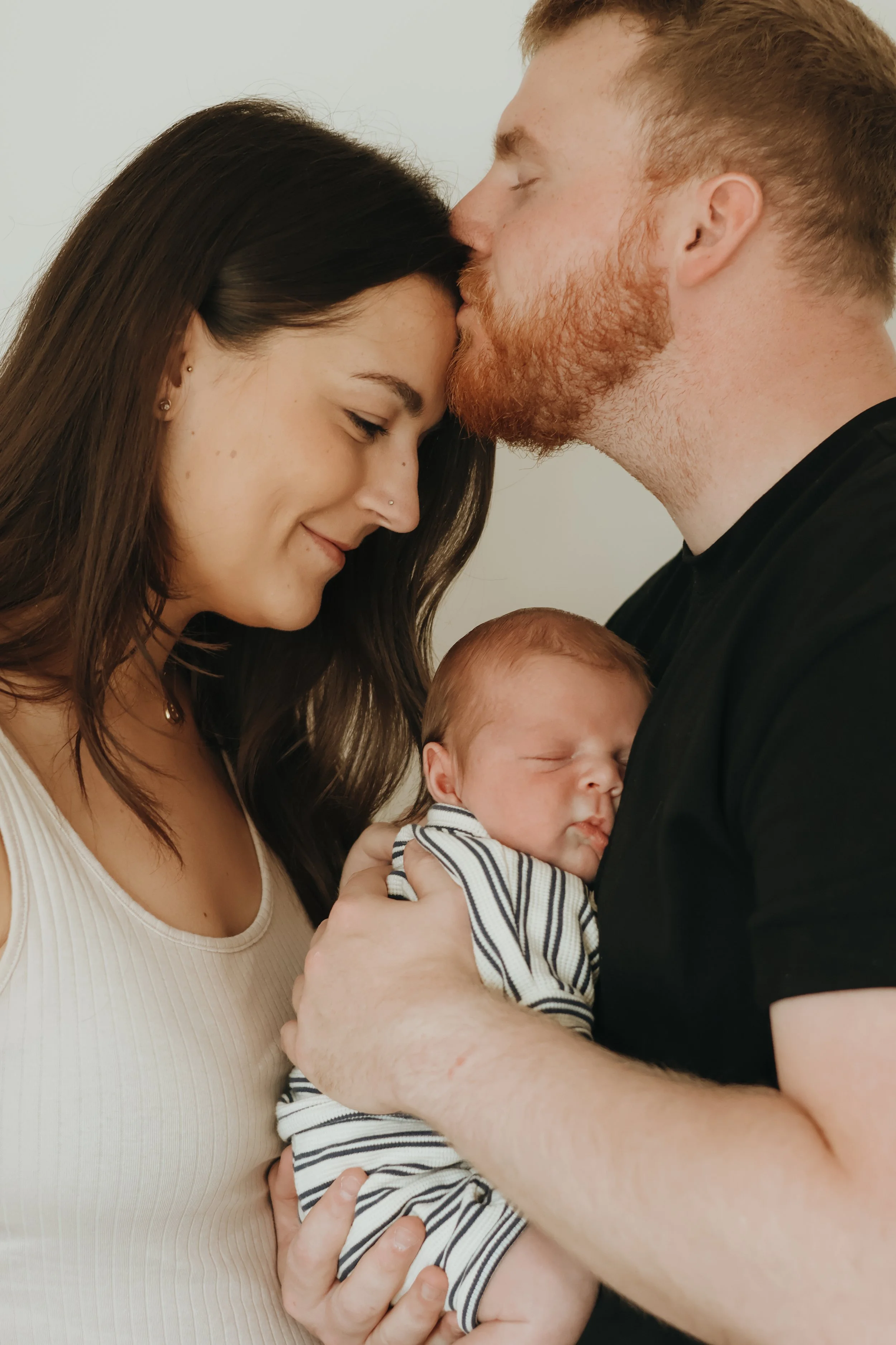 A family of three sharing a tender moment. The mother, with long dark hair, is smiling with her eyes closed as the father leans in to kiss her forehead. She is holding their sleeping newborn baby, who is dressed in a striped outfit. The father, with a red beard, has his eyes closed and is gently holding the baby.
