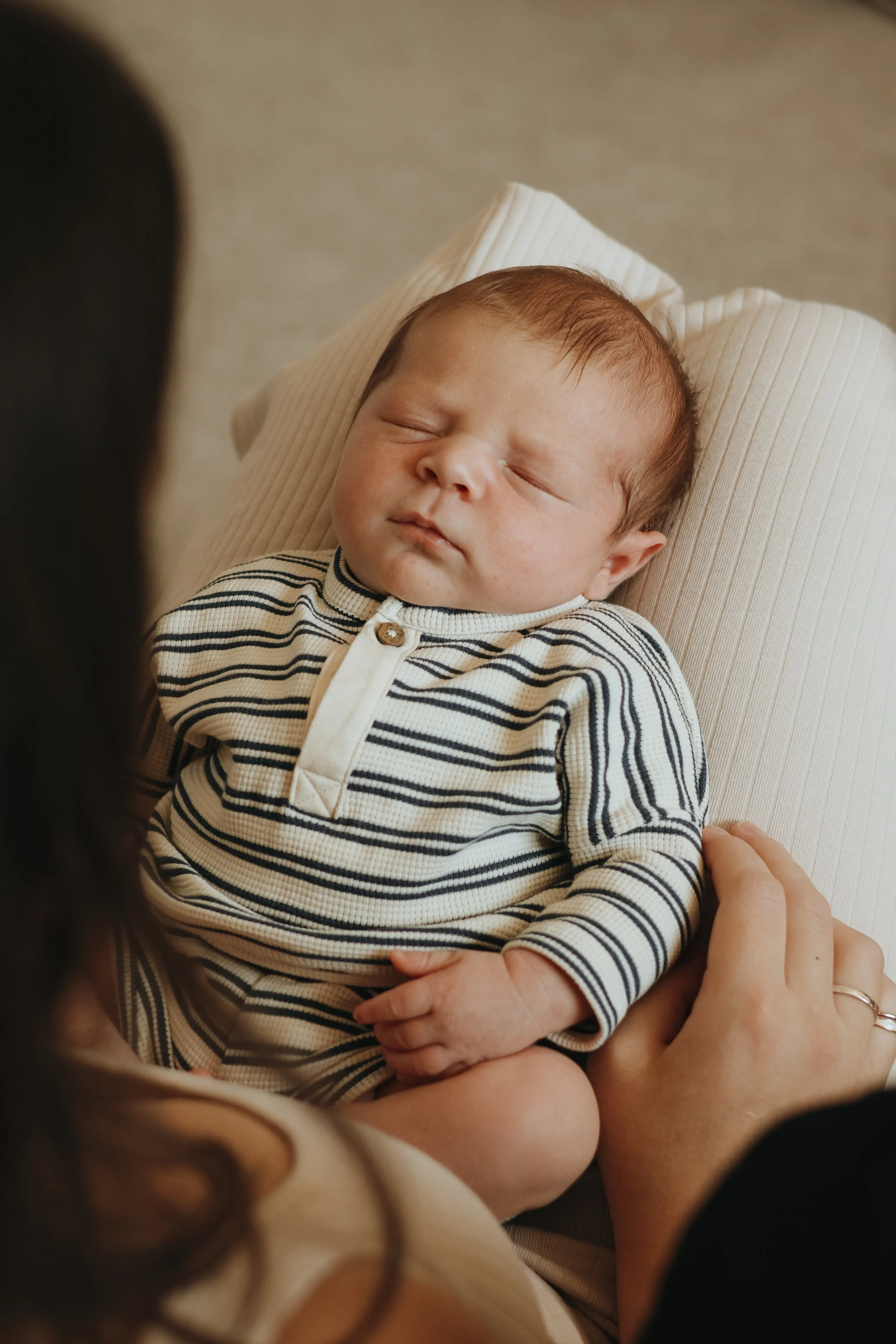 A sleeping baby with chubby cheeks, wearing a striped, long-sleeve shirt, lying on a pillow, while an adult's hand gently holds his arm.