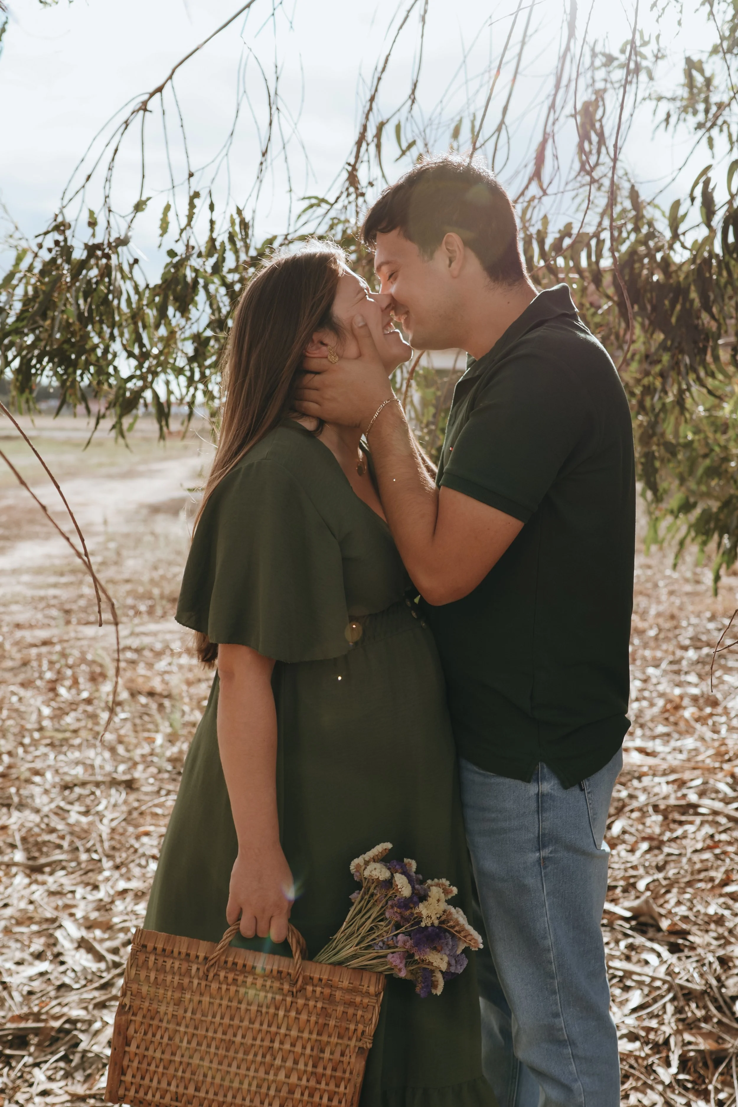 A couple embraces outdoors in a sunlit, wooded area, holding flowers and a basket, sharing an intimate moment.Maternity session, maternity photography, Portsmouth 