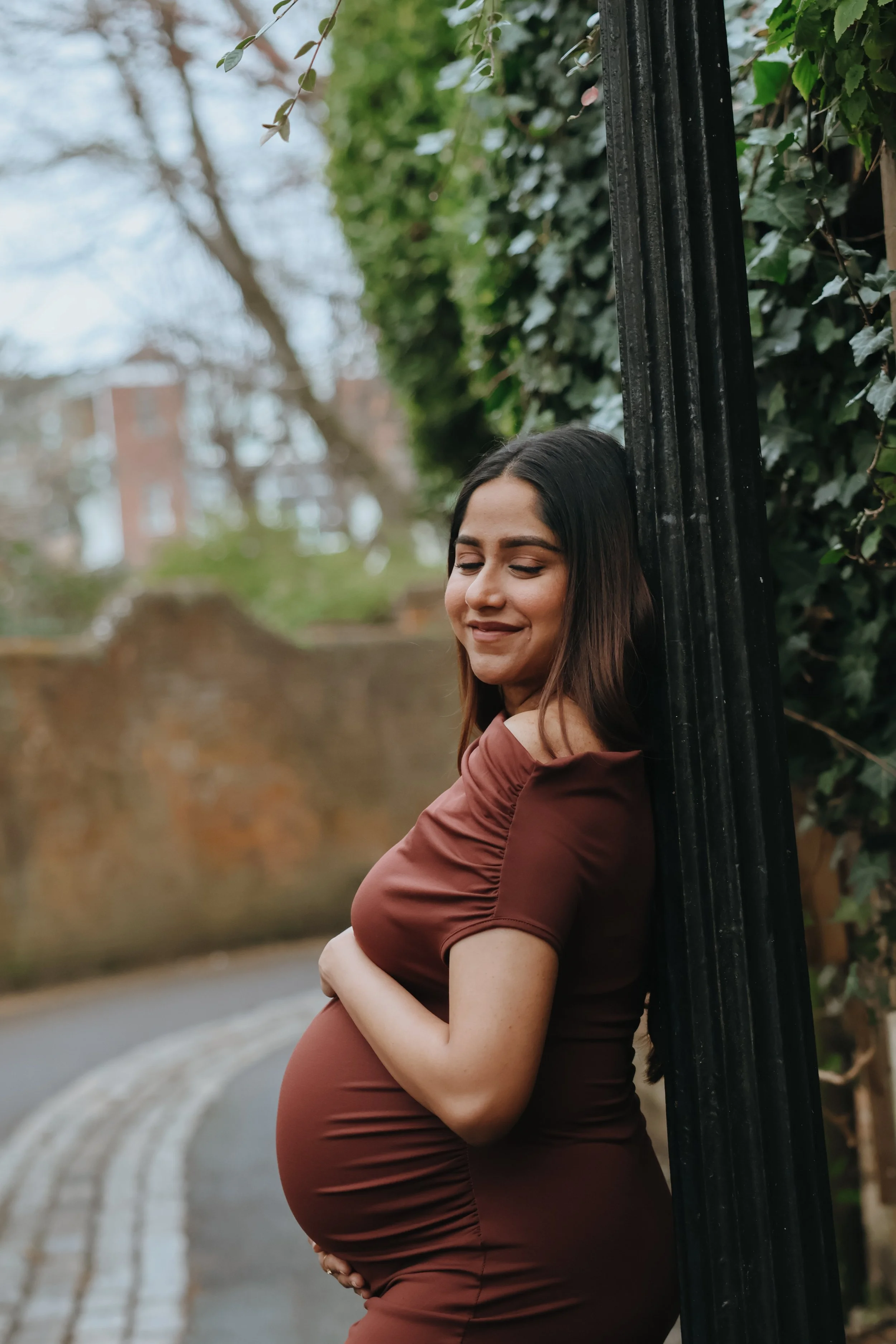 A pregnant woman with long dark hair, wearing a fitted brown dress, is leaning against a black pole outdoors, with her eyes closed and a smile. Maternity session, maternity photography, Portsmouth 
