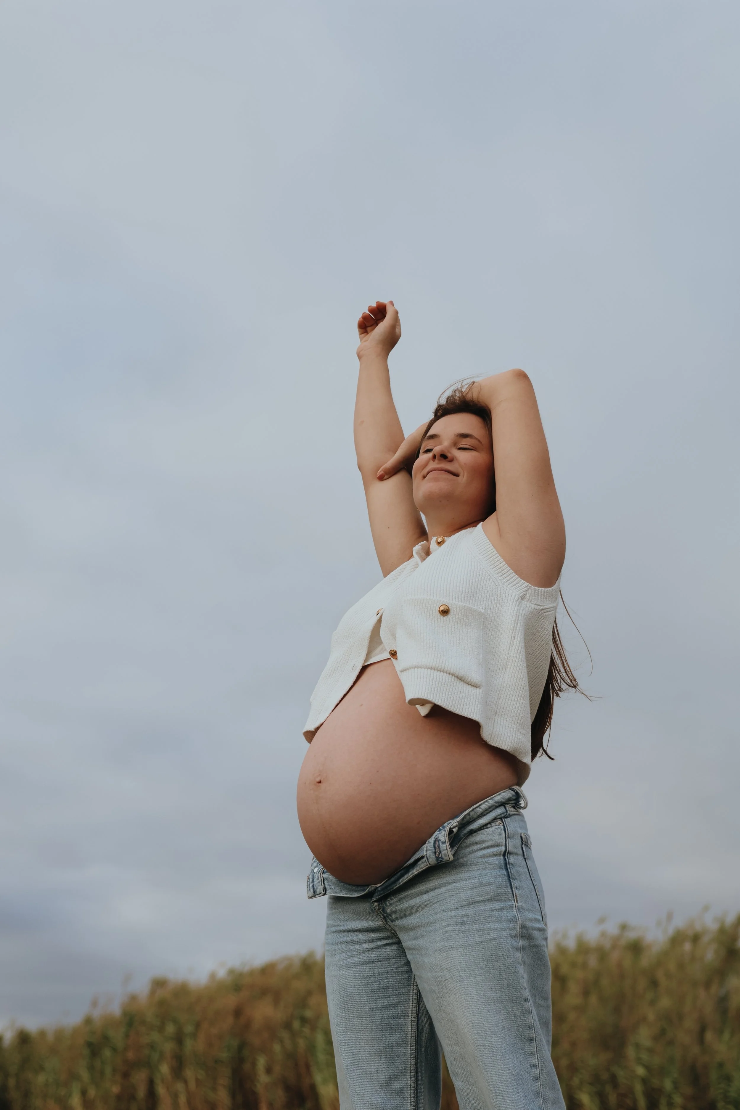 A pregnant woman standing outdoors, wearing a white sleeveless vest and jeans, with her arms raised and eyes closed, enjoying the moment.