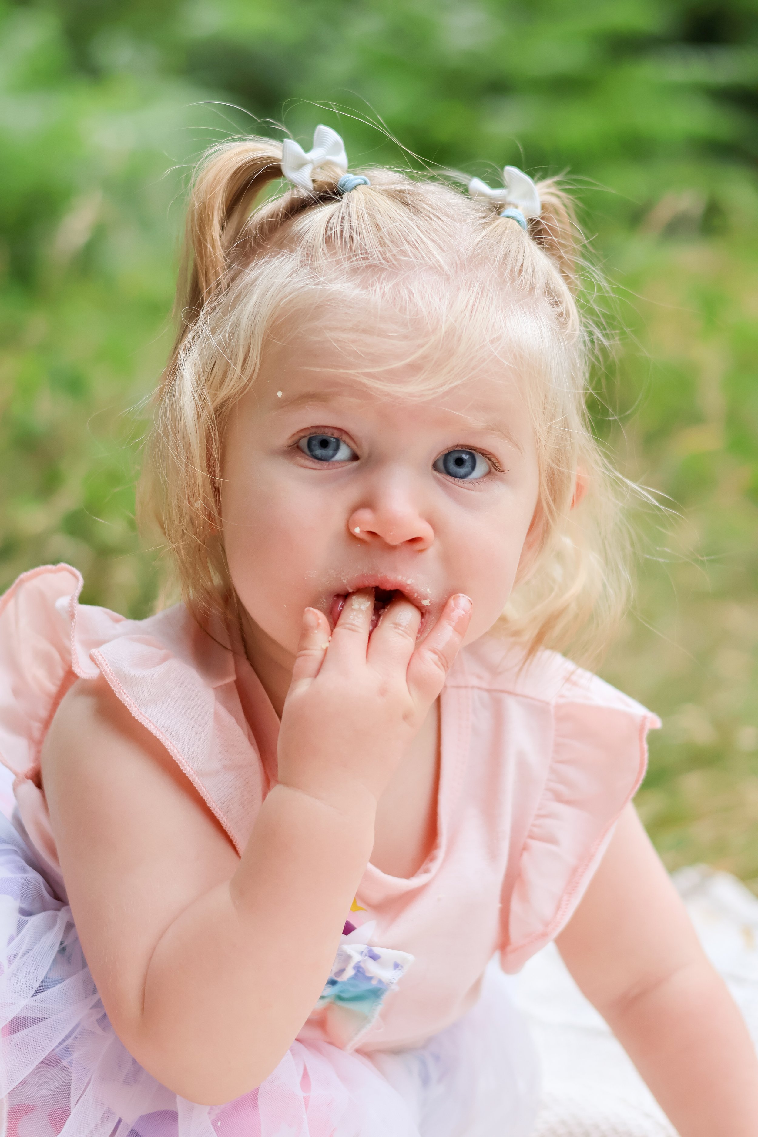 A young girl with blond hair in pigtails with white bows, wearing a pink dress, has her fingers in her mouth and is outdoors with green foliage in the background. cake smash, first birthday photography, southampton 