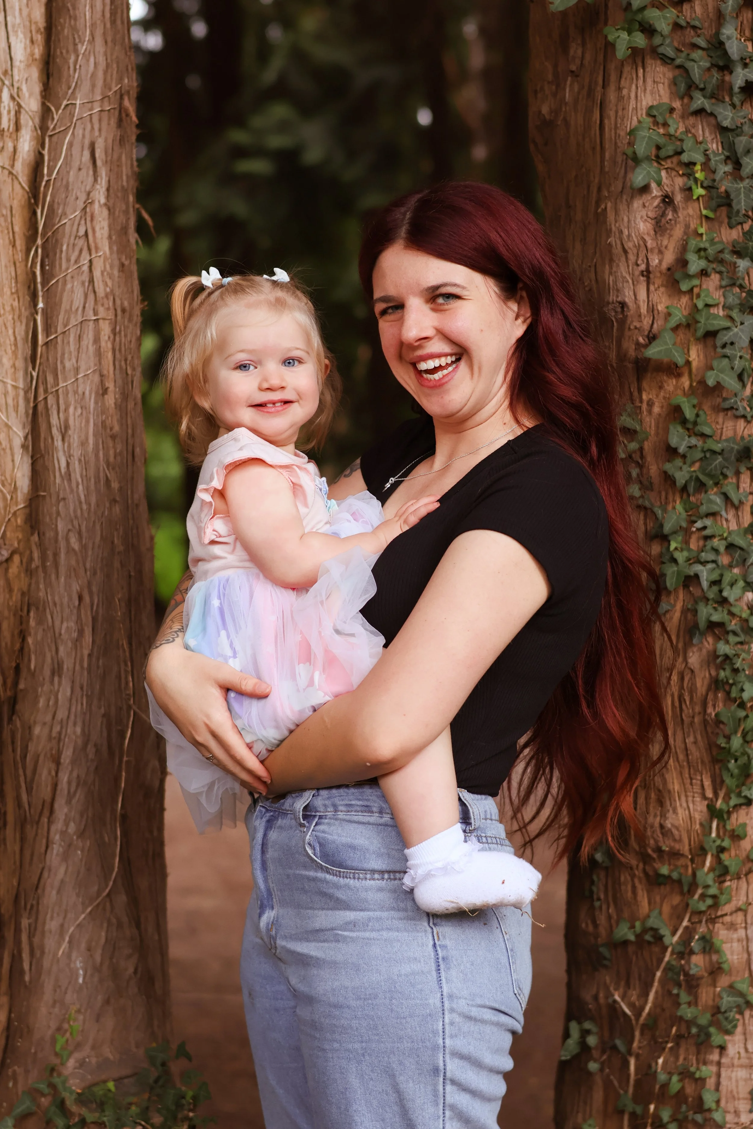 A woman with long red hair smiling and holding a young girl with blonde hair, dressed in a pastel dress, in a forest setting among trees.