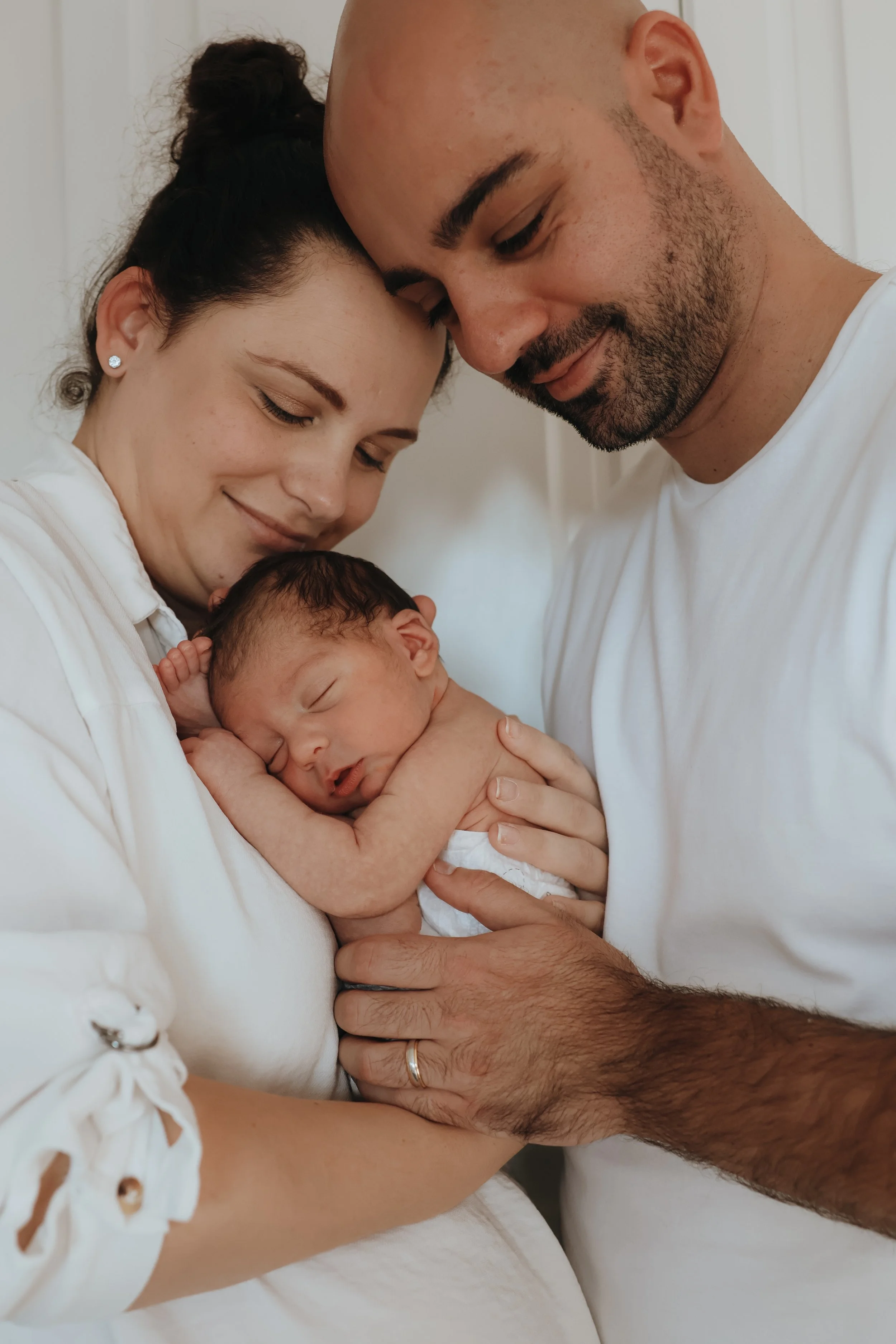 A mother and father holding their newborn baby together, all smiling gently, indoors with a white background. Family photography, newborn photography Bournemouth 