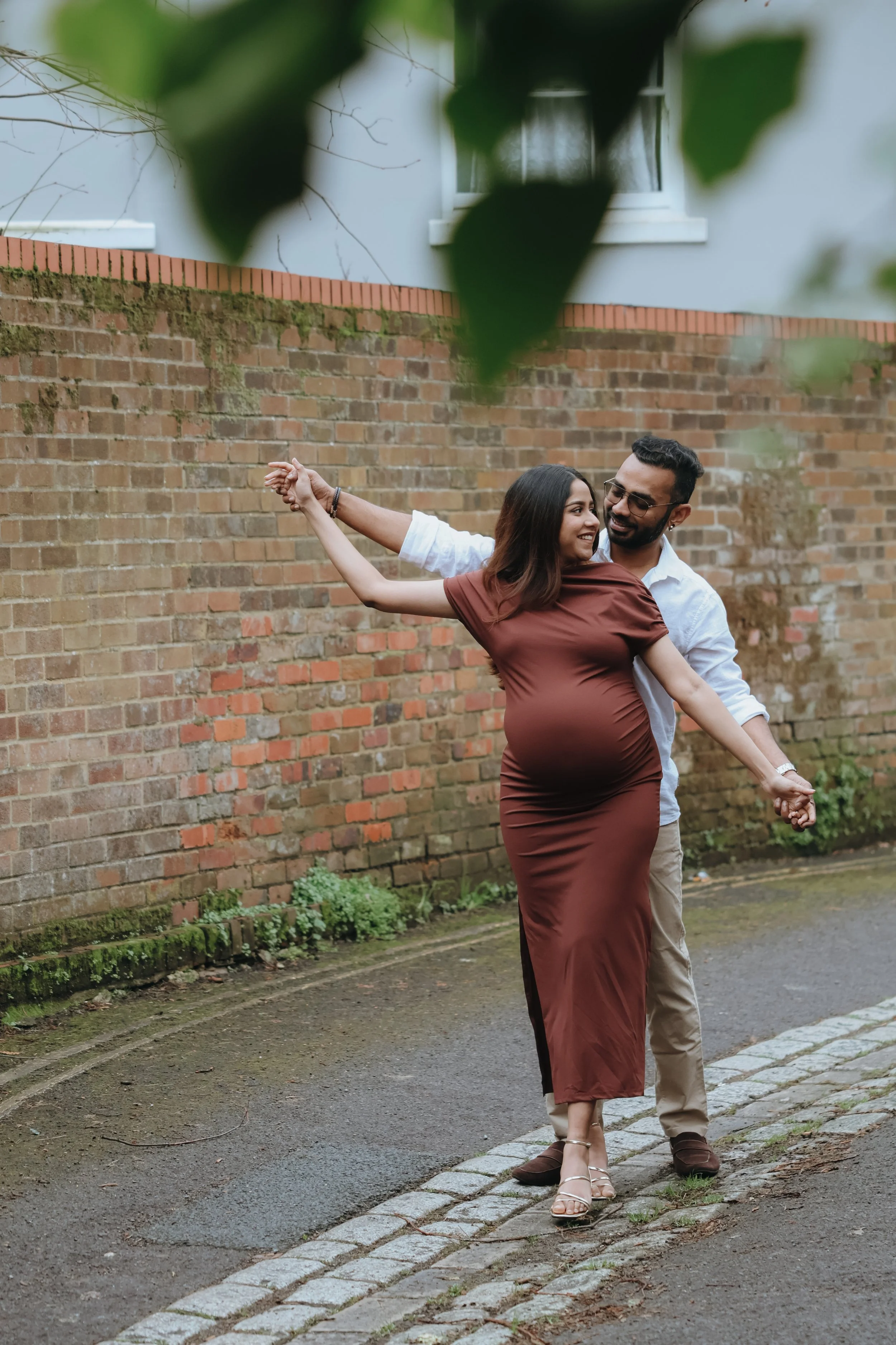 A couple dancing outdoors on a sidewalk with a brick wall in the background, the woman wearing a long brown dress and the man in a white shirt and beige pants. Maternity session, maternity photography, Portsmouth 