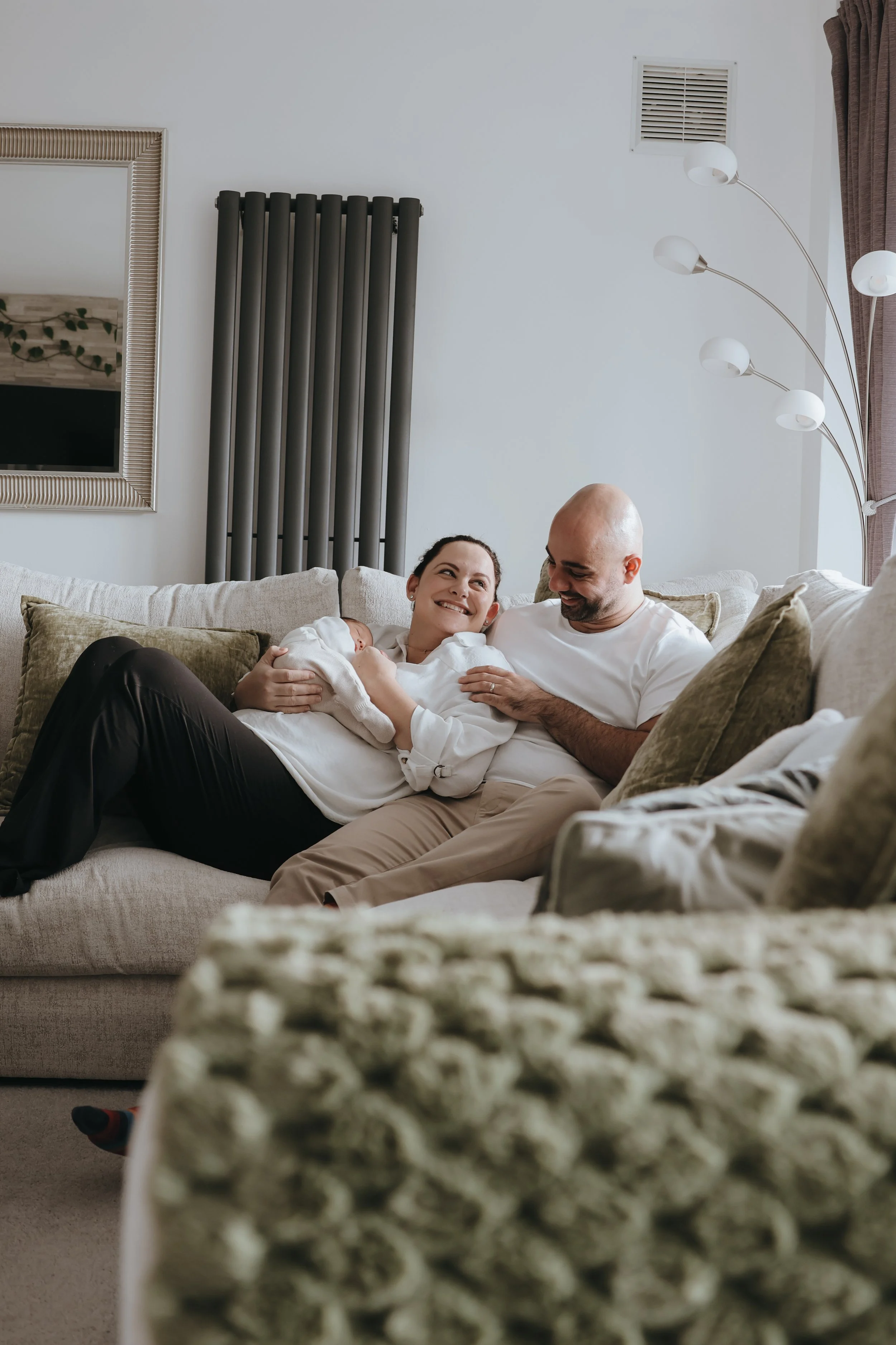 A family sitting on a beige couch in a living room, with the mother holding a newborn, all smiling and interacting. Family, newborn photography bournemouth 