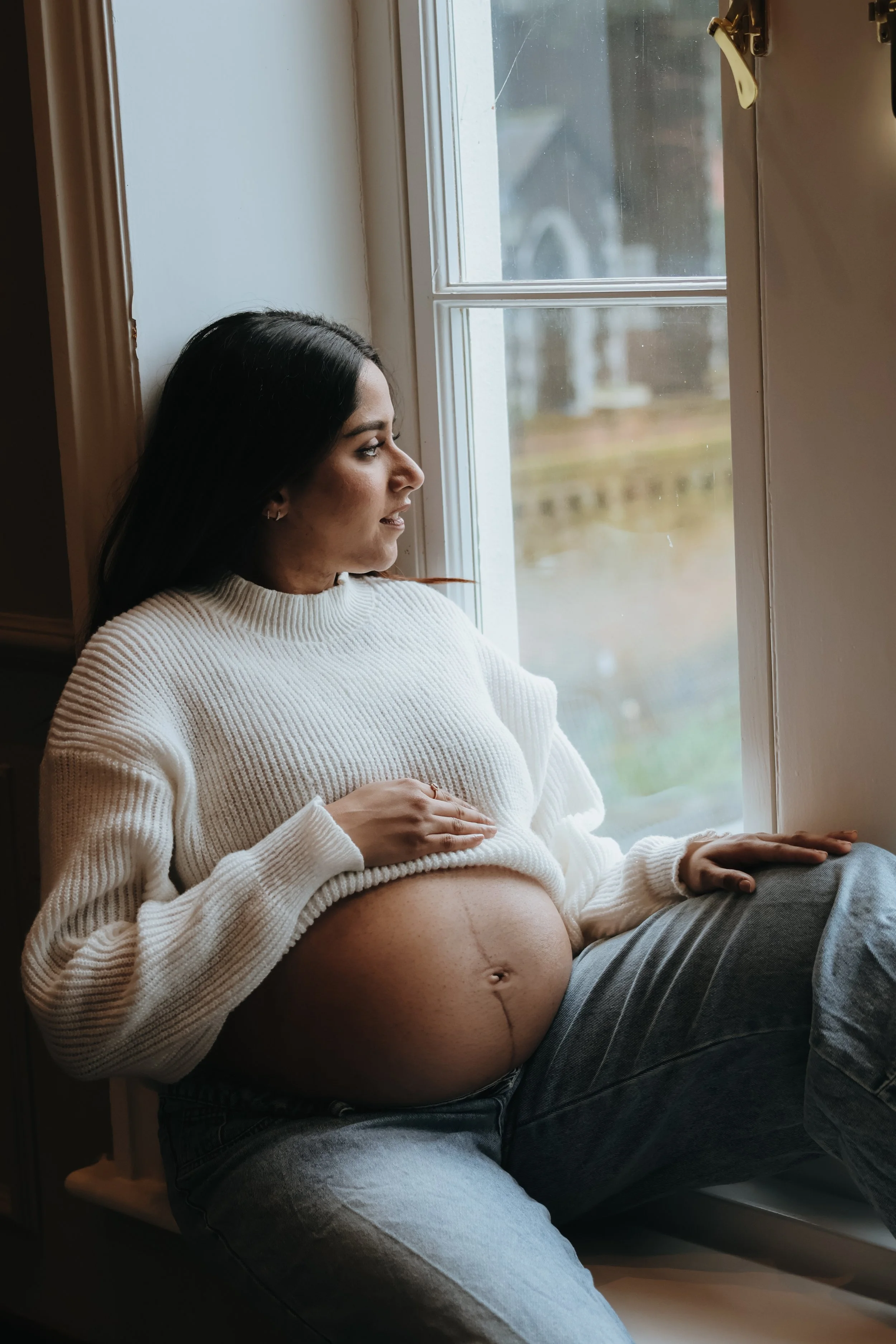 A pregnant woman sitting by a window, gazing outside, wearing a white sweater and gray pants. Maternity session, maternity photography, Portsmouth 
