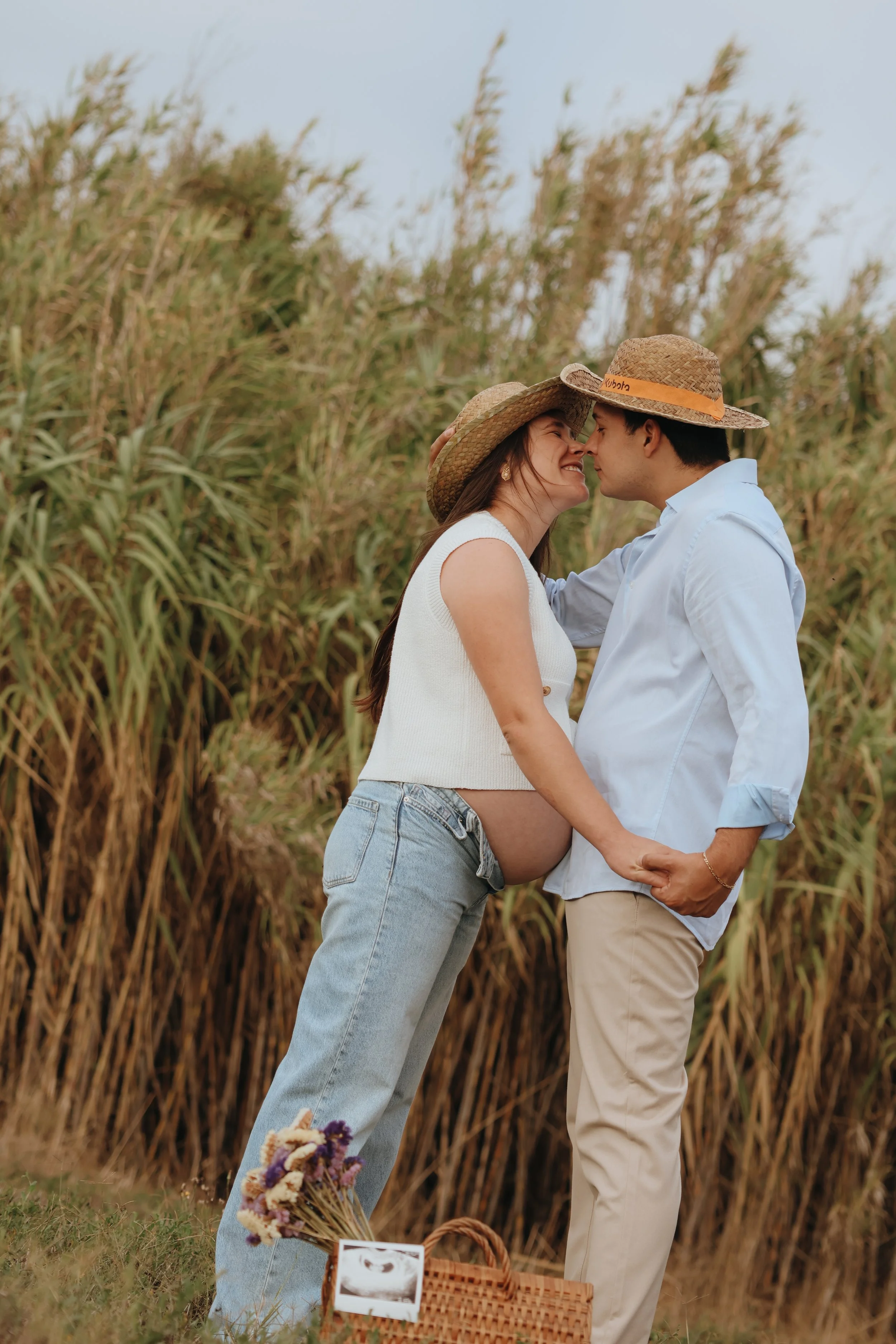 A couple stands in a field with tall golden grass, nose to nose, smiling and holding hands. The woman is pregnant, wearing a white sleeveless top, light-wash jeans, and a wide-brimmed hat. The man is dressed in a light blue shirt, beige pants, and a straw hat. A picnic basket with flowers and a photograph is in the foreground.