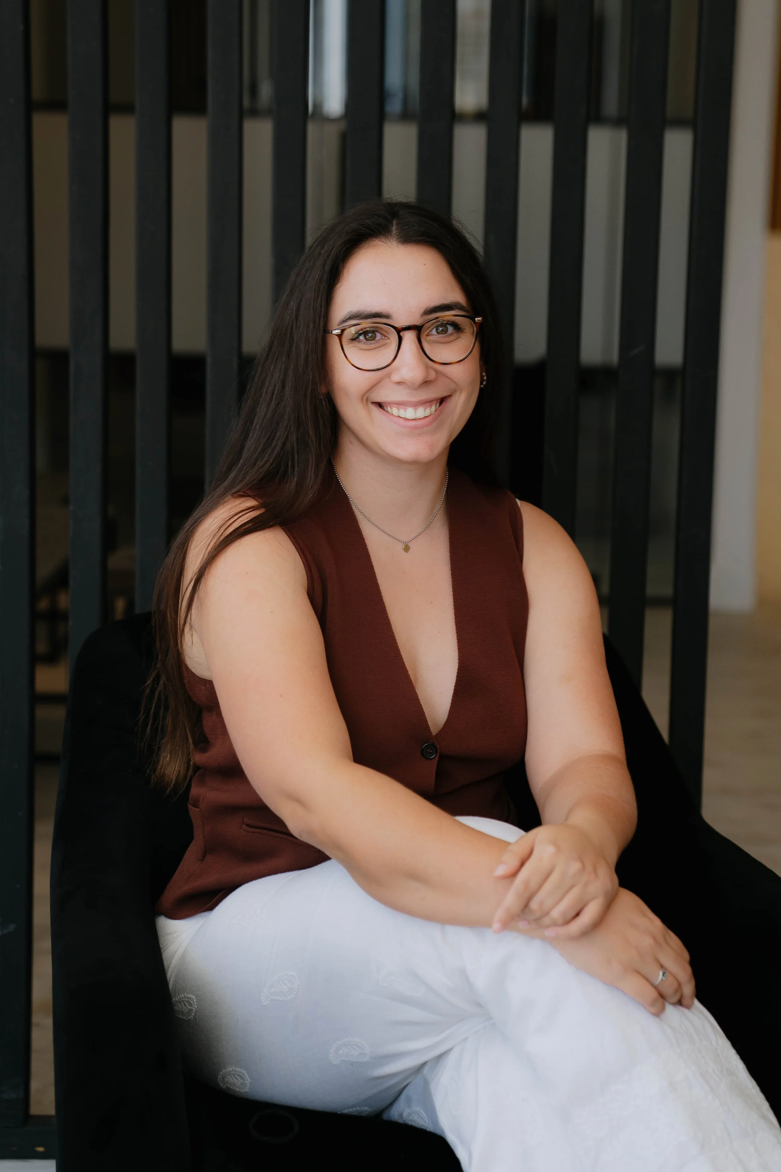 A woman with long dark hair, glasses, and a smile, sitting on a black chair in front of a dark railing, wearing a sleeveless brown top and white pants.