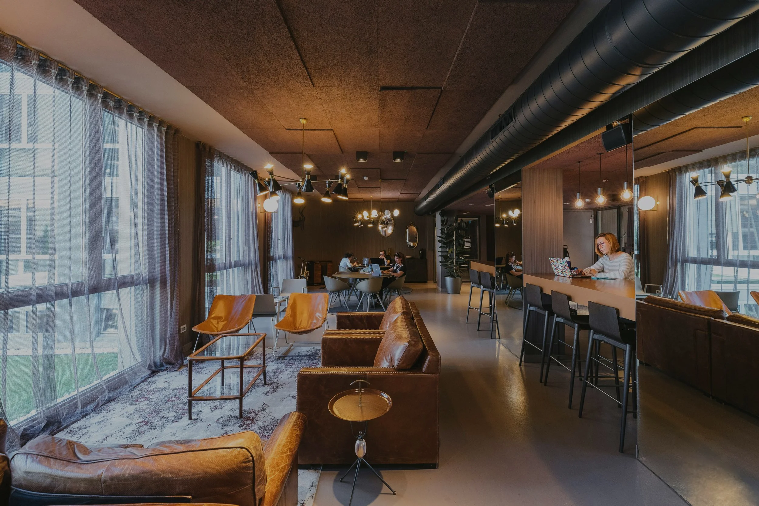 Modern office lounge with leather couches, wooden tables, and chairs. People working on laptops at a long high table. Large windows with sheer curtains and industrial lighting overhead.