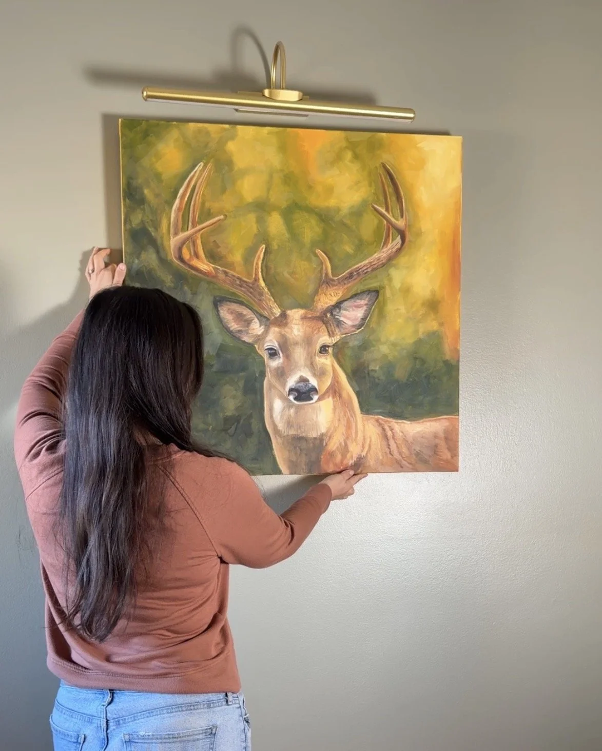 Woman hanging a landscape painting of a deer with large antlers on a wall.
