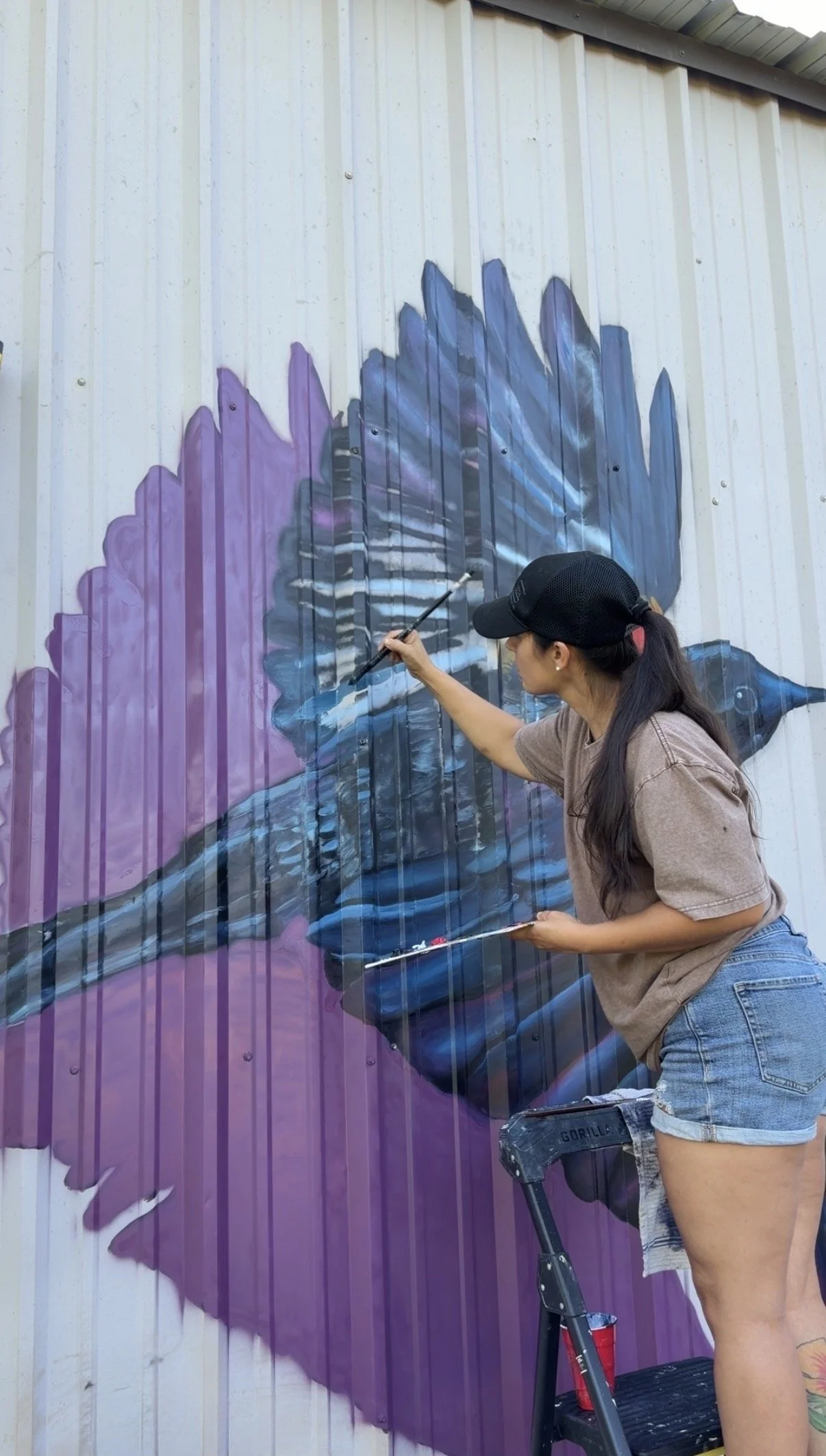 A woman painting a mural on a vertical metal wall, painting a purple and blue whale in a realistic style.