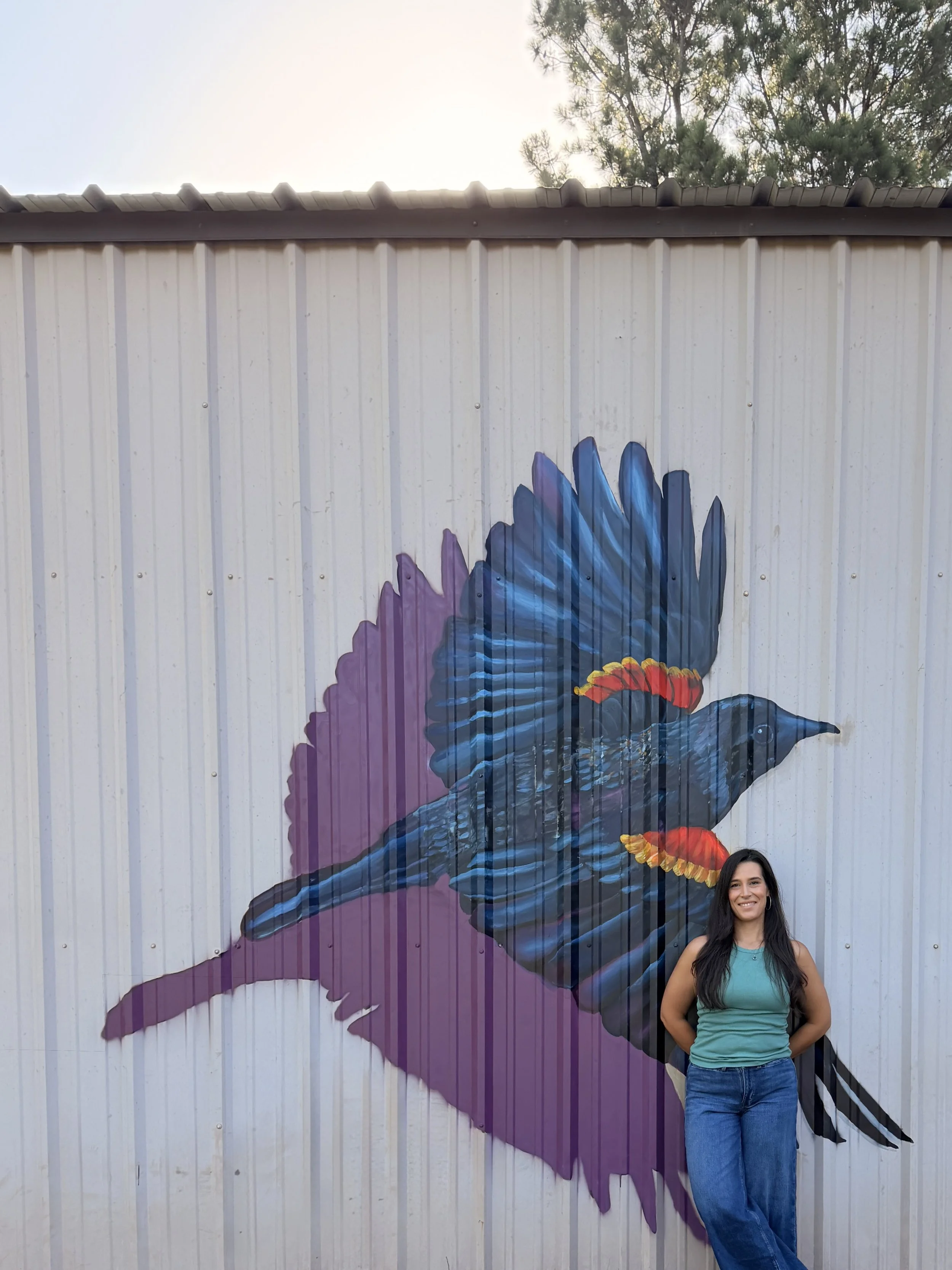 A woman standing in front of a painted mural of a bird on a metal wall, with trees and sky visible in the background.