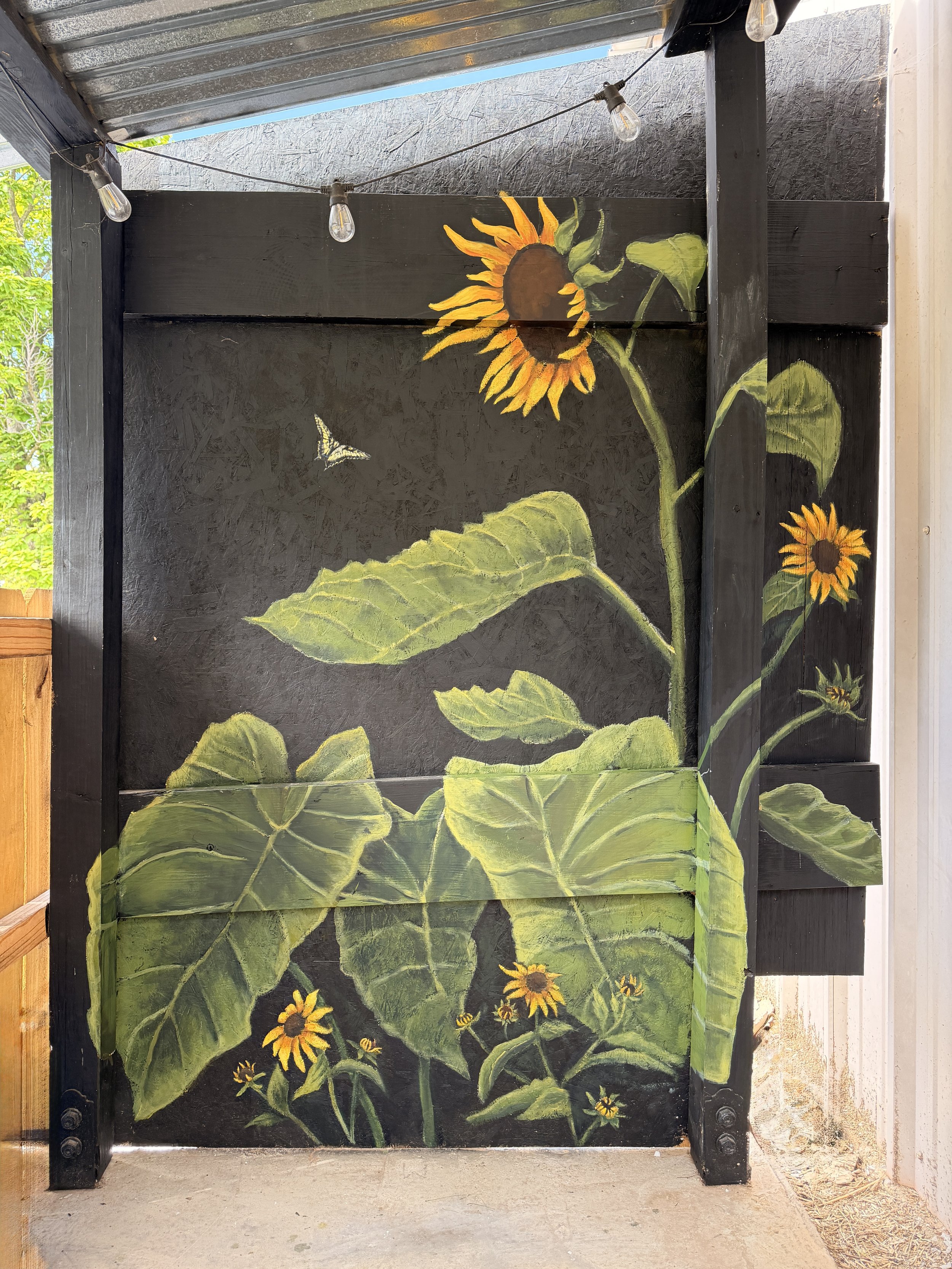 A painted mural of sunflowers and a butterfly on a black wooden surface, with string lights hanging above.