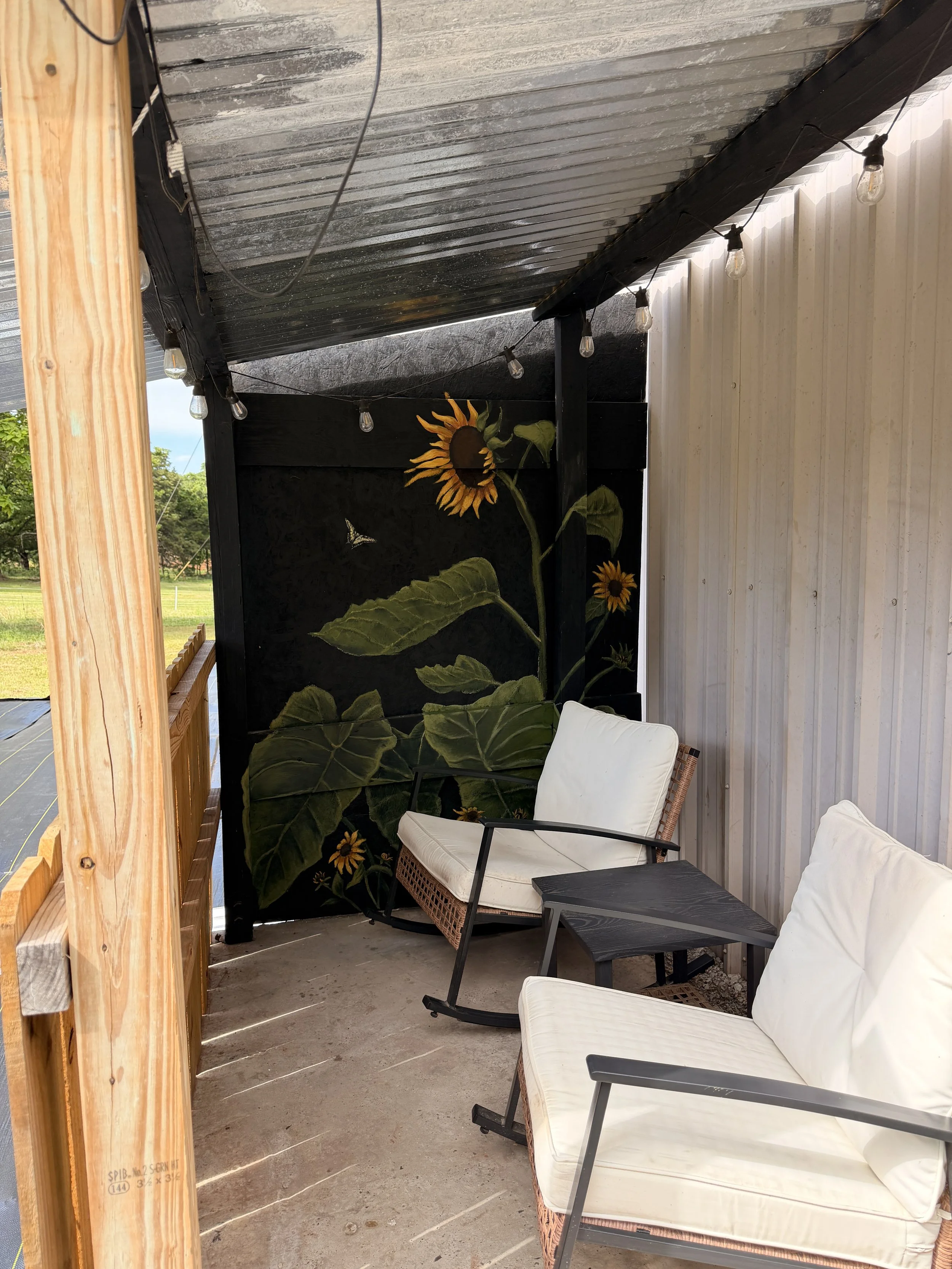 Outdoor patio area with black and white walls, string lights, a sunflower mural, wicker chairs with cushions, and a small black table.