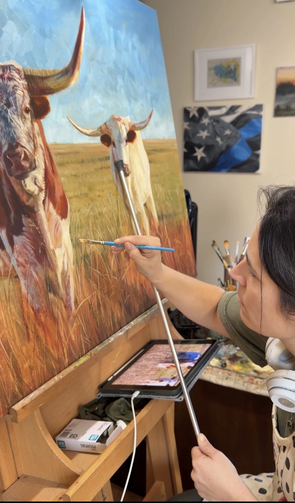 A woman painting a landscape with cows on a canvas on an easel. The woman is using a long-handled brush and has a palette on her lap. The studio has several framed artworks on the wall and a wooden shelf with art supplies and a box of PopShins branded tissues.