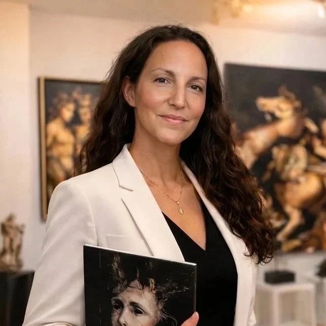 A woman with long curly brown hair, wearing a white blazer over a black top, holding a black folder with a picture of a child's face on it, standing in an art gallery with visible paintings of horses and trophies in the background.