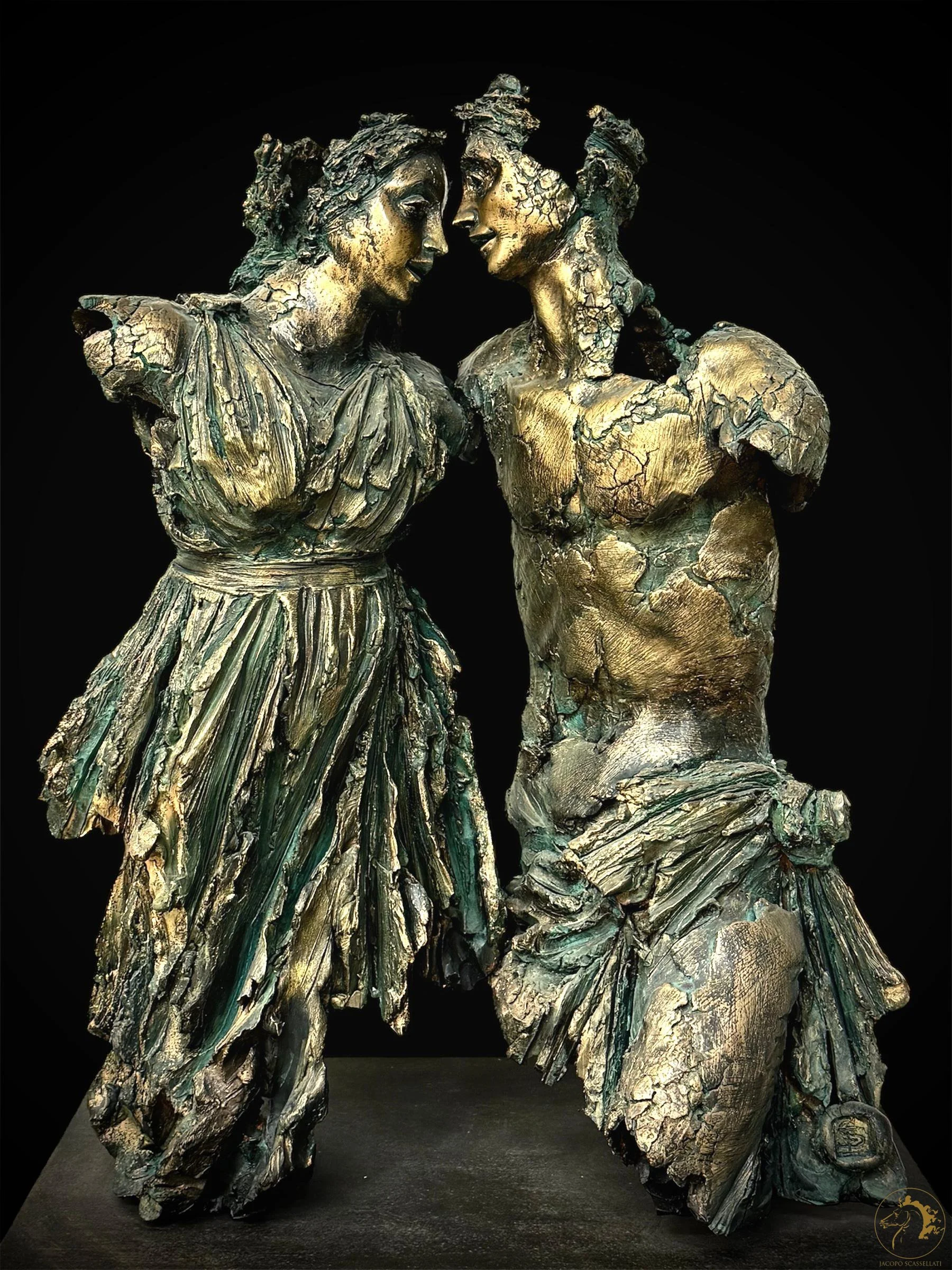 A sculpture of two women in ancient Greek style, facing each other with their foreheads touching, made of a textured, metallic material with gold and greenish patina, set against a black background.