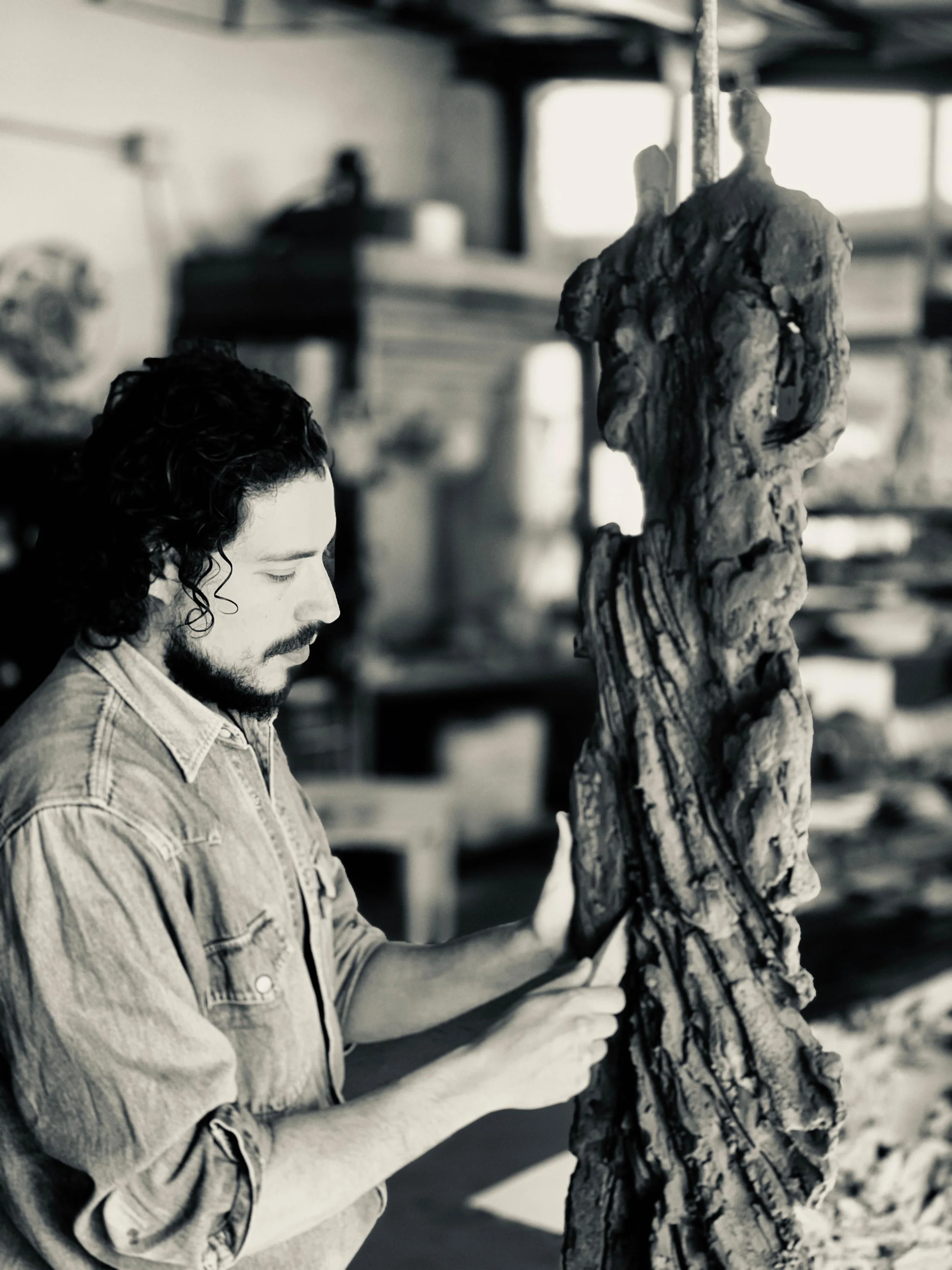 A man with curly hair and a beard is in a workshop, touching a large, textured sculptural piece made of wood or clay, with shelves and tools in the background.