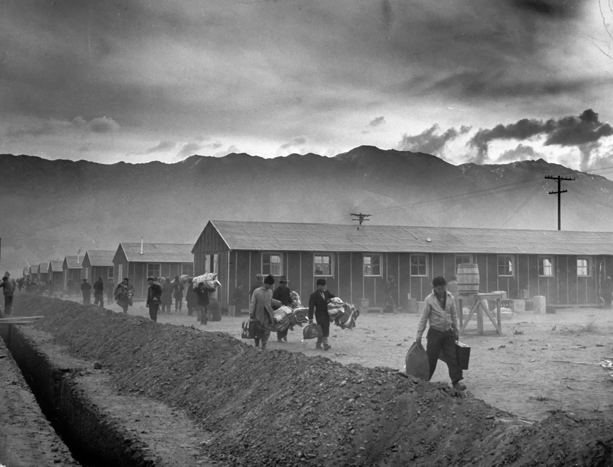 Black and white photo of a rural area with mountains in the background, people walking with bags, and buildings with sloped roofs in the foreground.