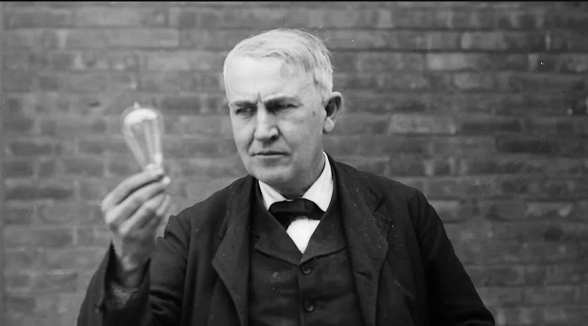 A man with light-colored hair and a serious expression holding a light bulb in his right hand while looking at it, standing in front of a brick wall.