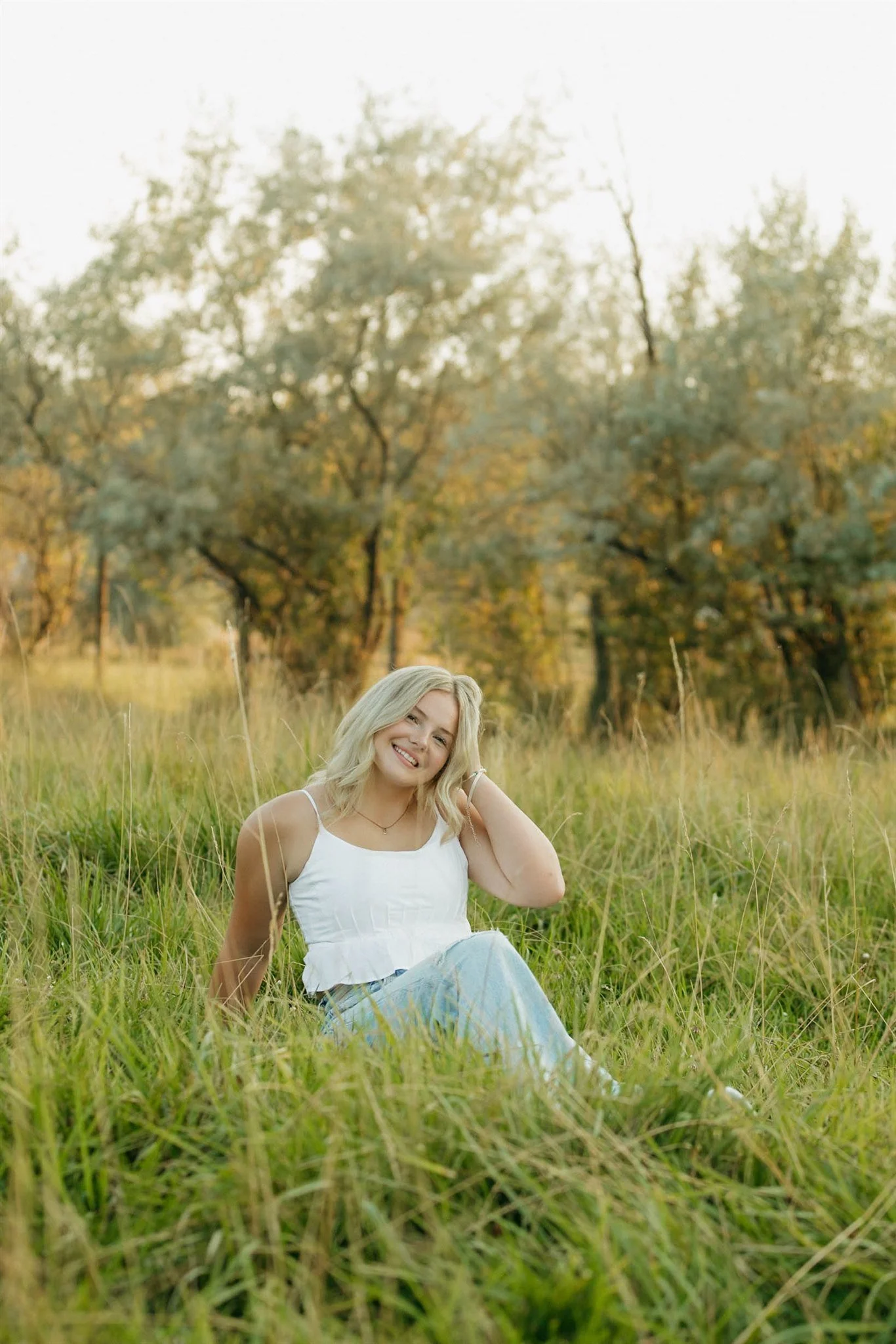 A young woman with blonde hair sitting in tall grass in a field during autumn, smiling and touching her hair, with trees in the background.
