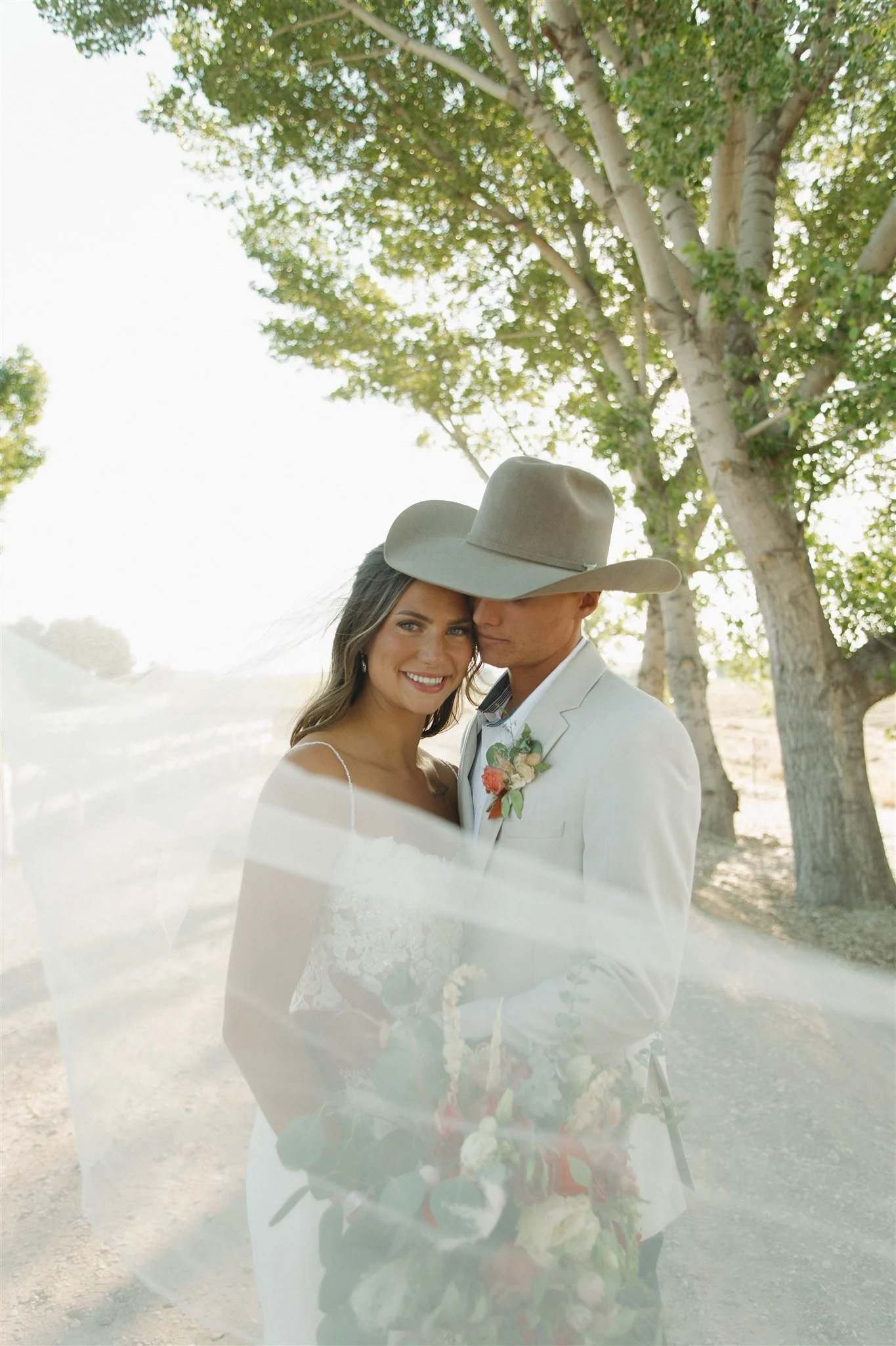 A bride and groom outdoors with tall trees in the background, sharing an intimate moment. The bride is smiling, and the groom is wearing a cowboy hat. The bride has a white dress with lace details, and the groom is in a light-colored suit with a boutonniere. Soft sunlight filters through the trees.