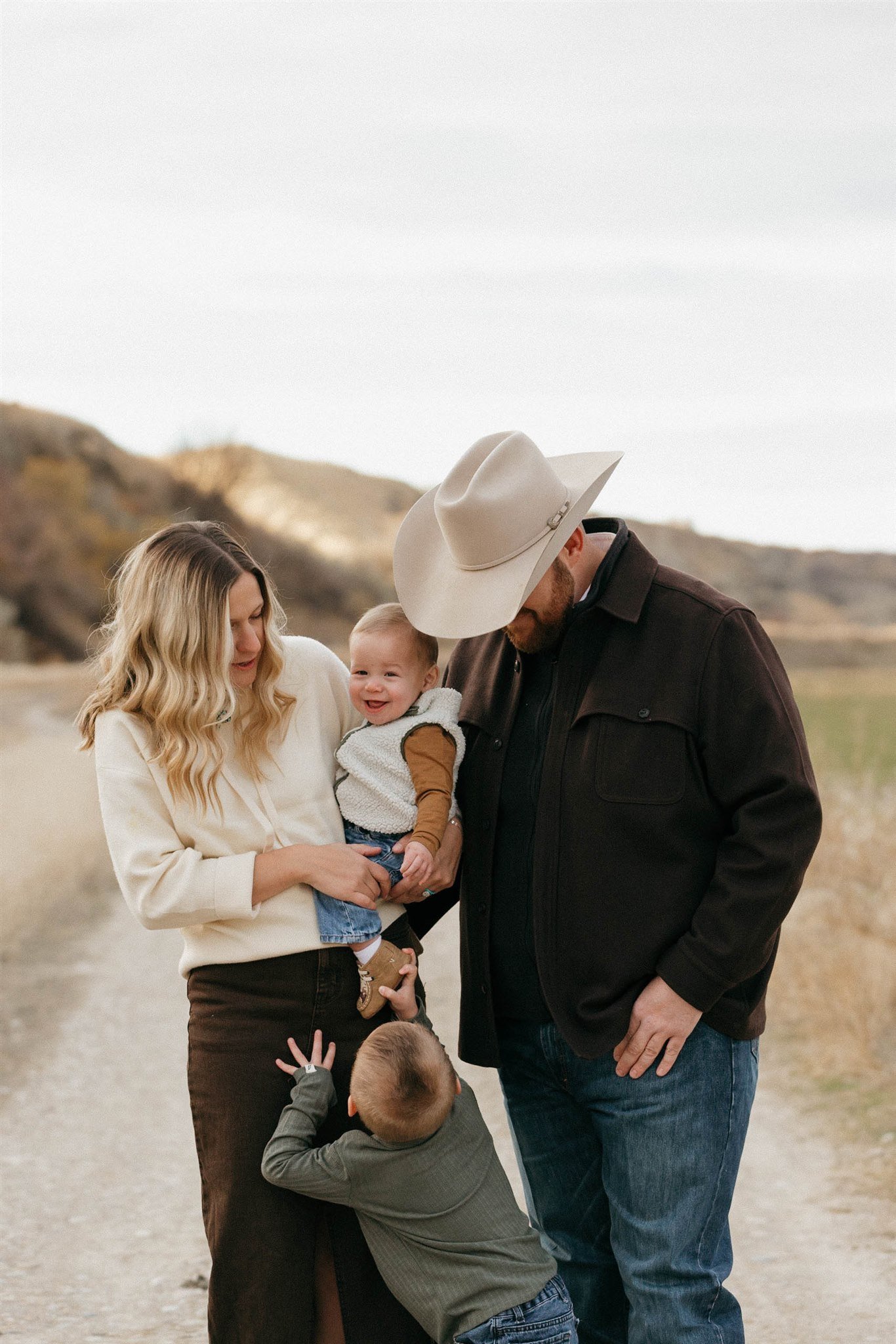 A family of four enjoying time outdoors on a dirt path with hills in the background, the woman holding a smiling baby and a young boy standing at her feet looking up, the man wearing a cowboy hat standing next to them.