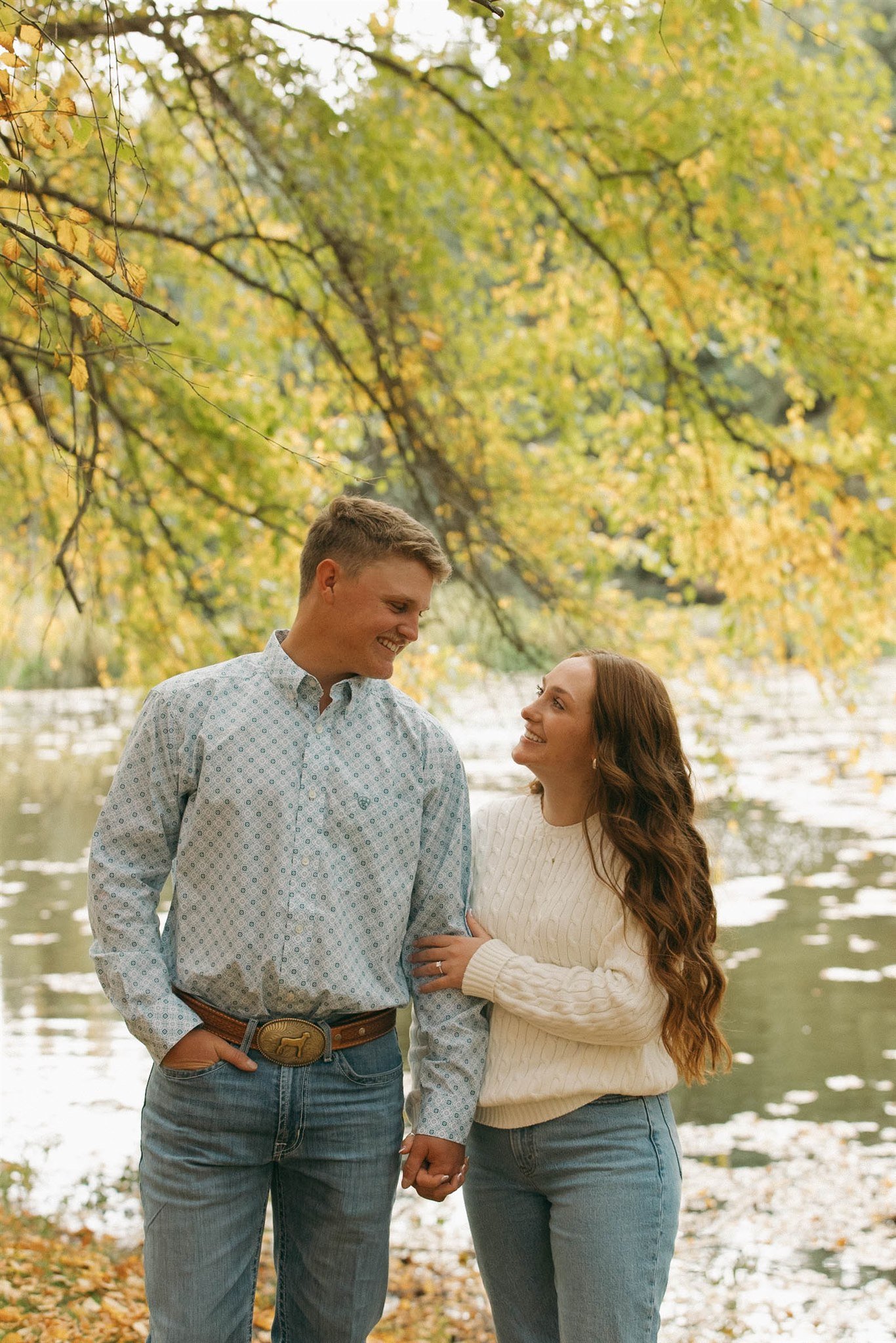 A couple holding hands and smiling at each other by a river surrounded by trees with fall foliage.