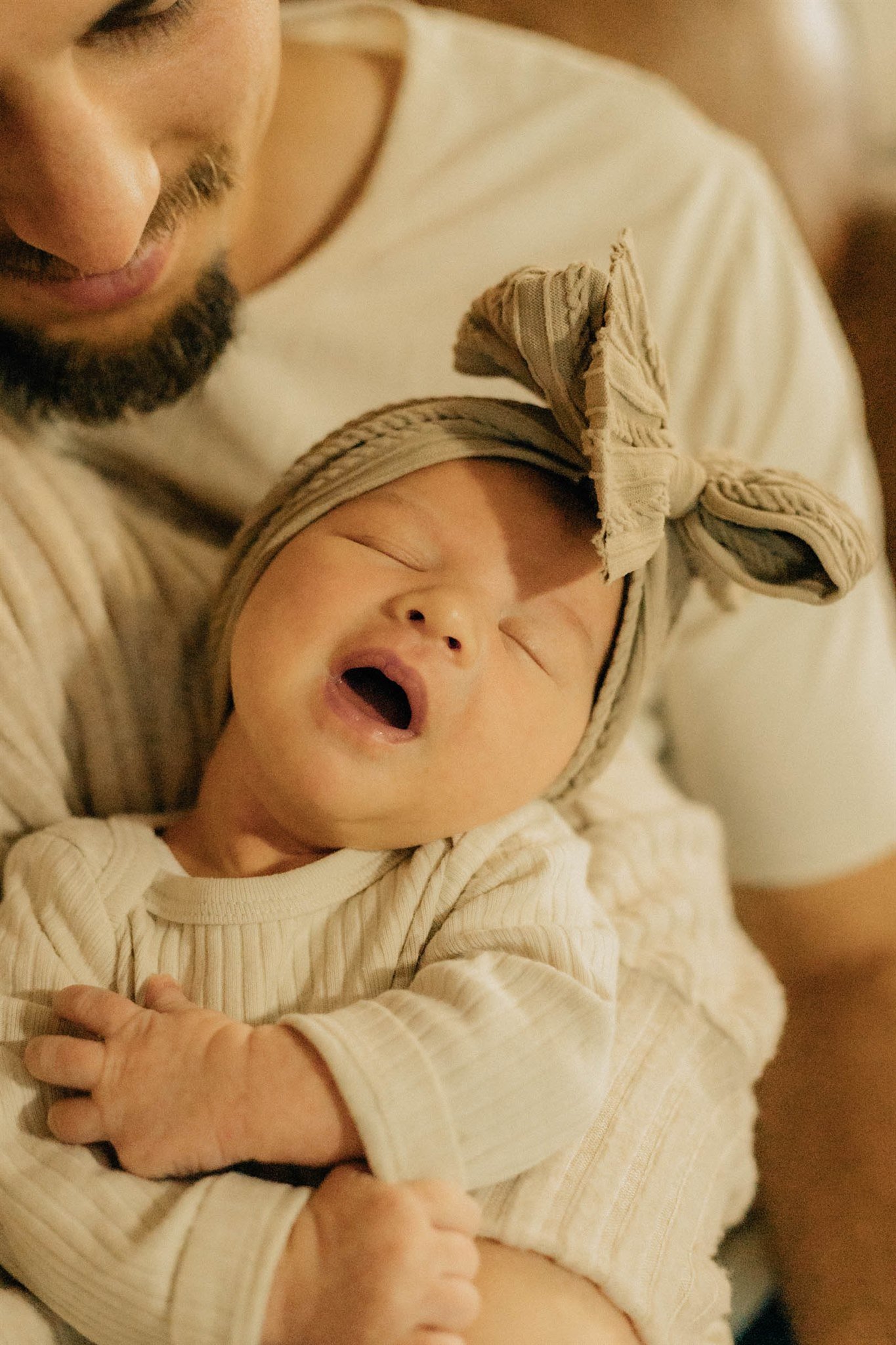 A man holding a young child, who is yawning and wearing a beige striped outfit and a matching head wrap with a bow.
