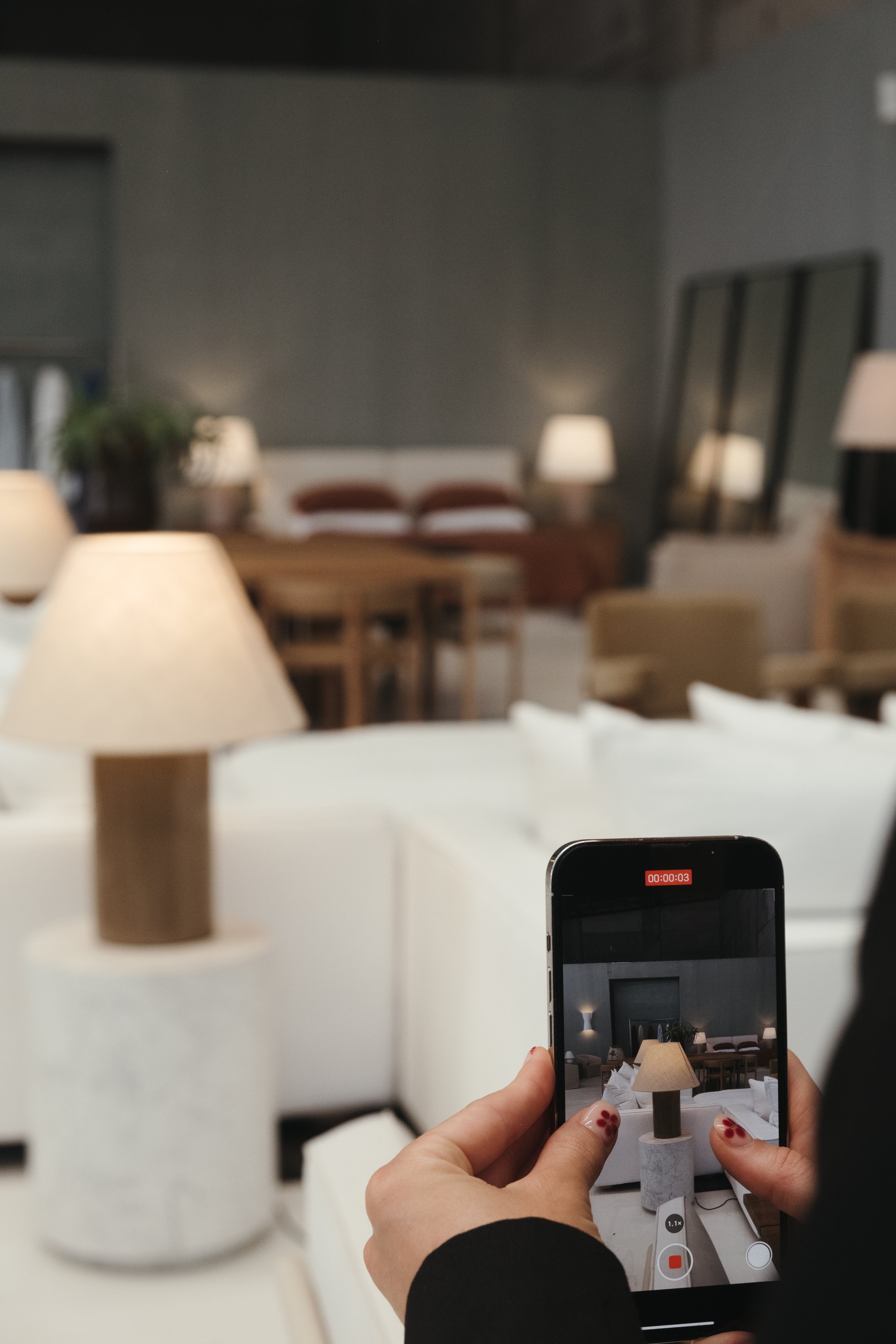 Person recording a video of a lamp on a side table in a modern, well-lit living room with sofas, a dining table, and wall mirrors.