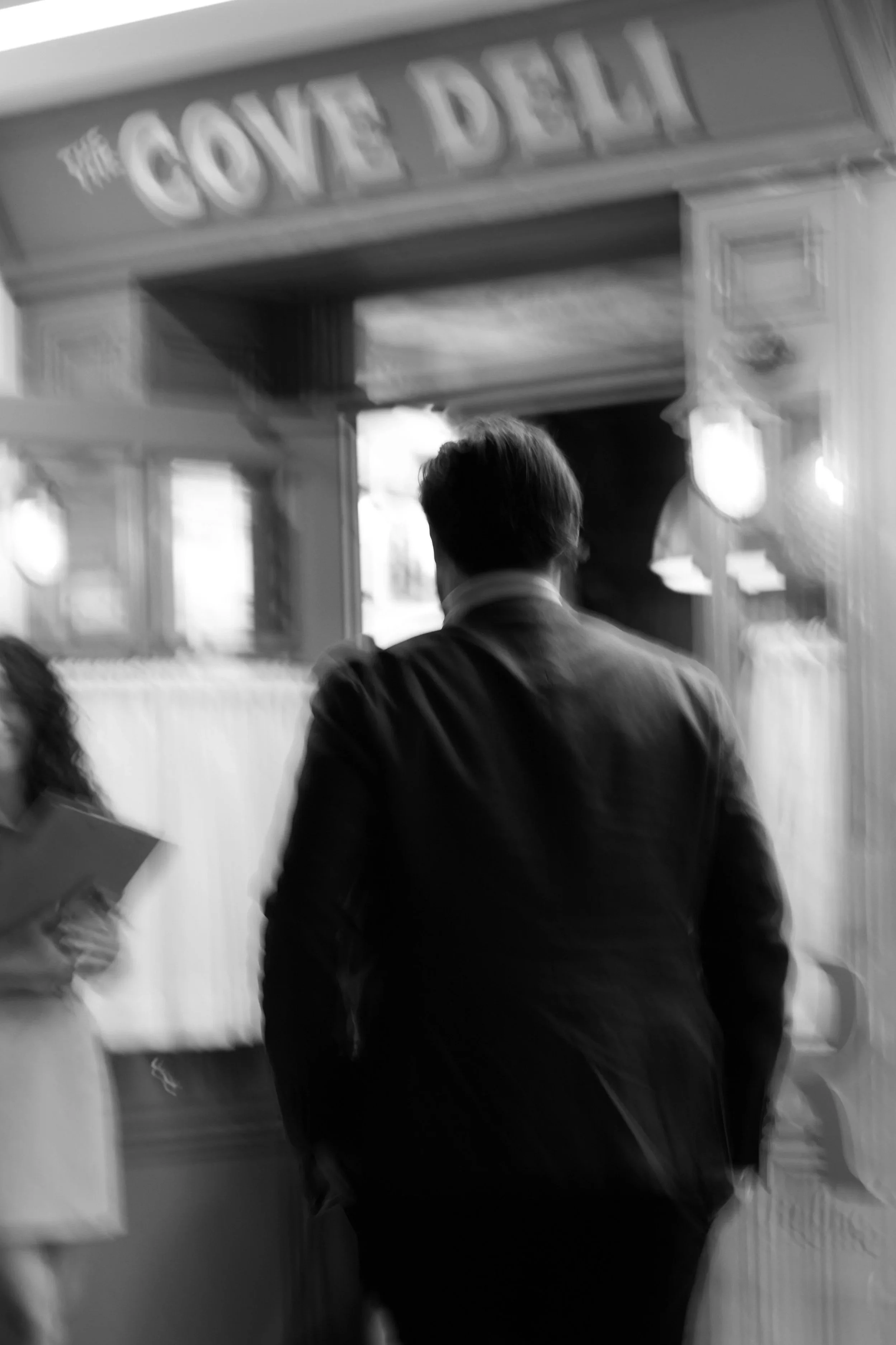 Back view of a man in a suit standing inside a bakery or deli with a sign above that reads 'The GONE DELI.'