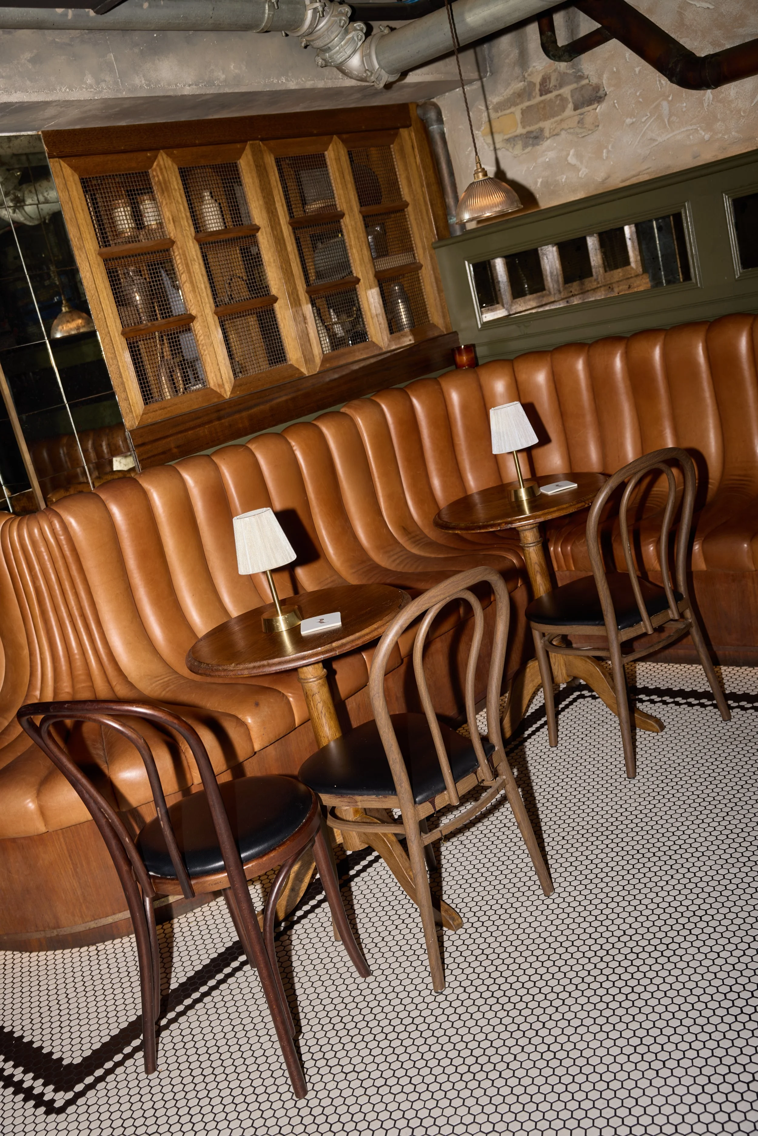 Interior of a cozy cafe with brown leather banquette seating, small round wooden tables, and wooden chairs with black cushioned seats. There are small white lamps on the tables, a wood-paneled wall behind the seating, and a rustic brick wall with industrial pipes.
