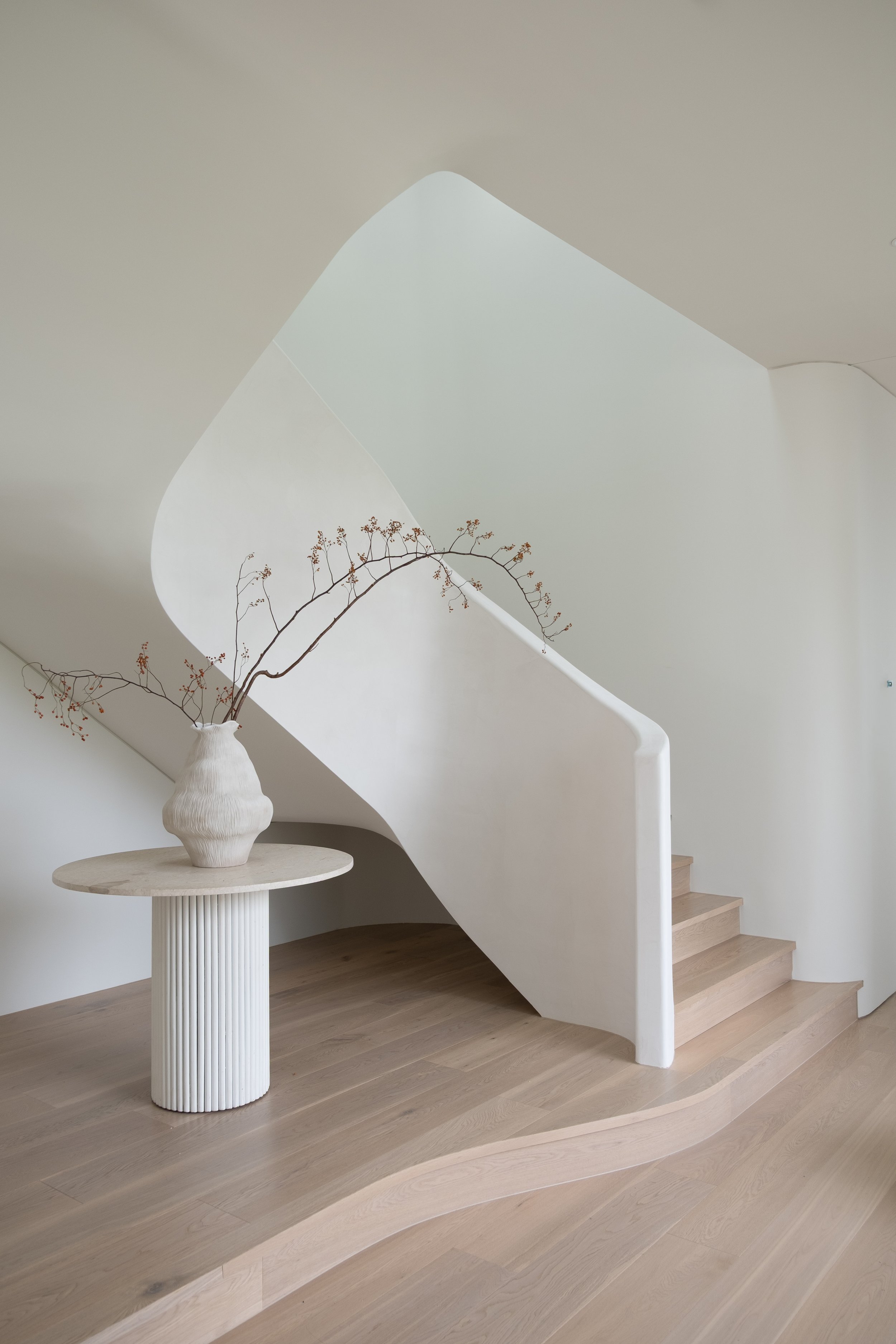 Minimalist interior featuring a curved staircase with wooden steps, a white sculptural handrail, a white textured side table, and a white ceramic vase with a dried branch.