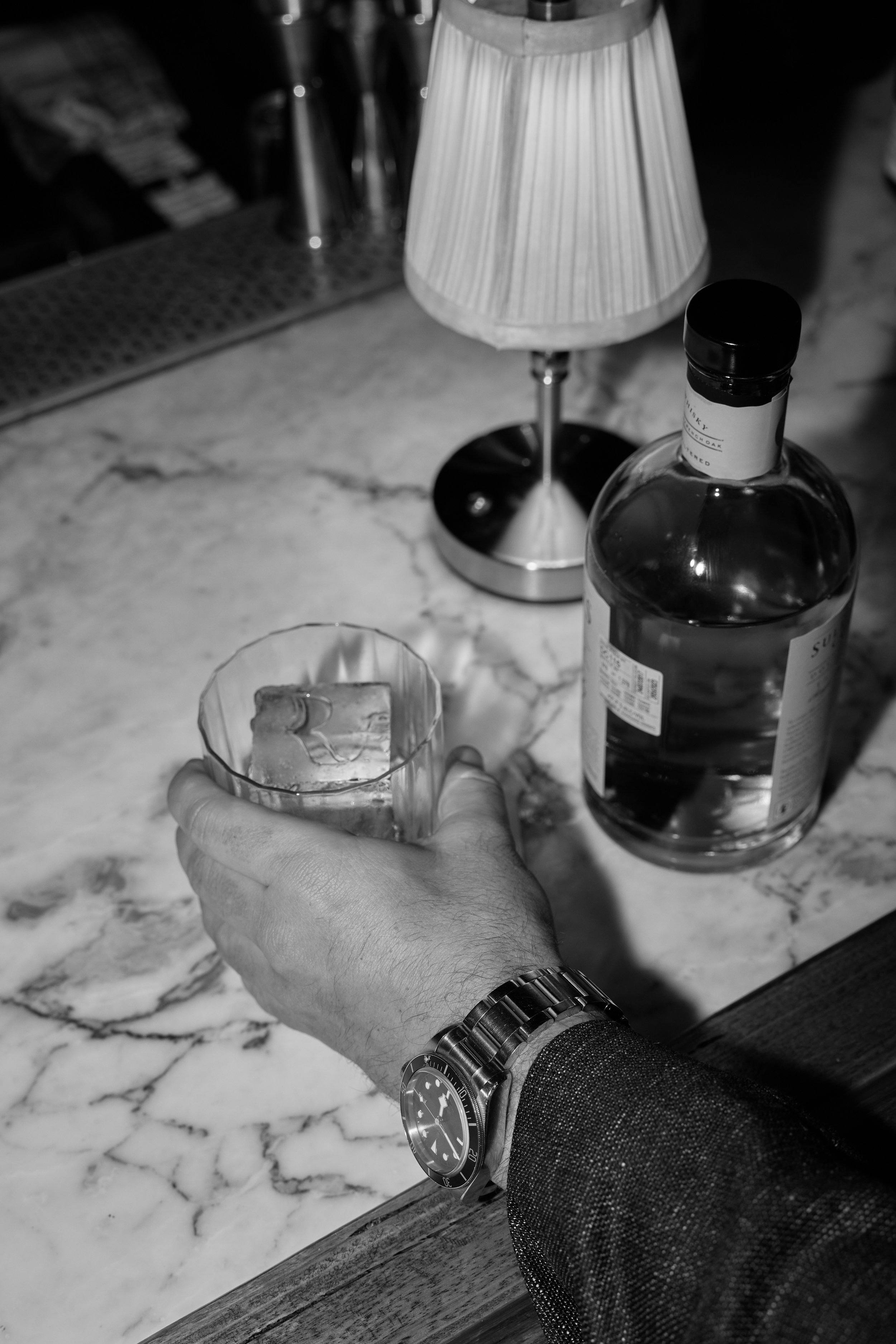 A person wearing a watch is holding a glass with a drink on a marble bar counter. A bottle of alcohol and a small lamp are also on the counter.