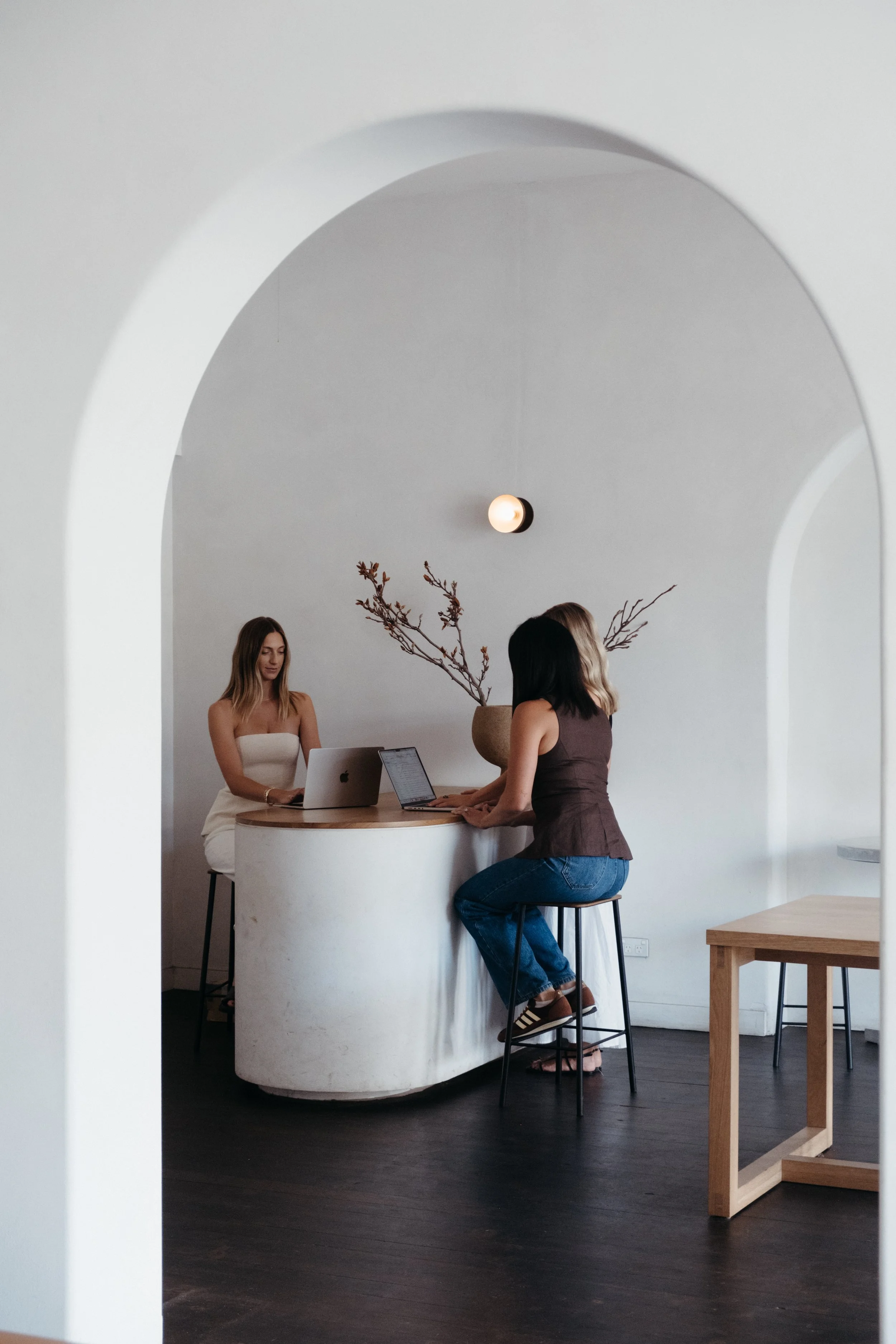 Two women are working on laptops at a white marble desk in a minimalistic room with white walls, a black ceiling light, and a large beige vase with branches as decor.