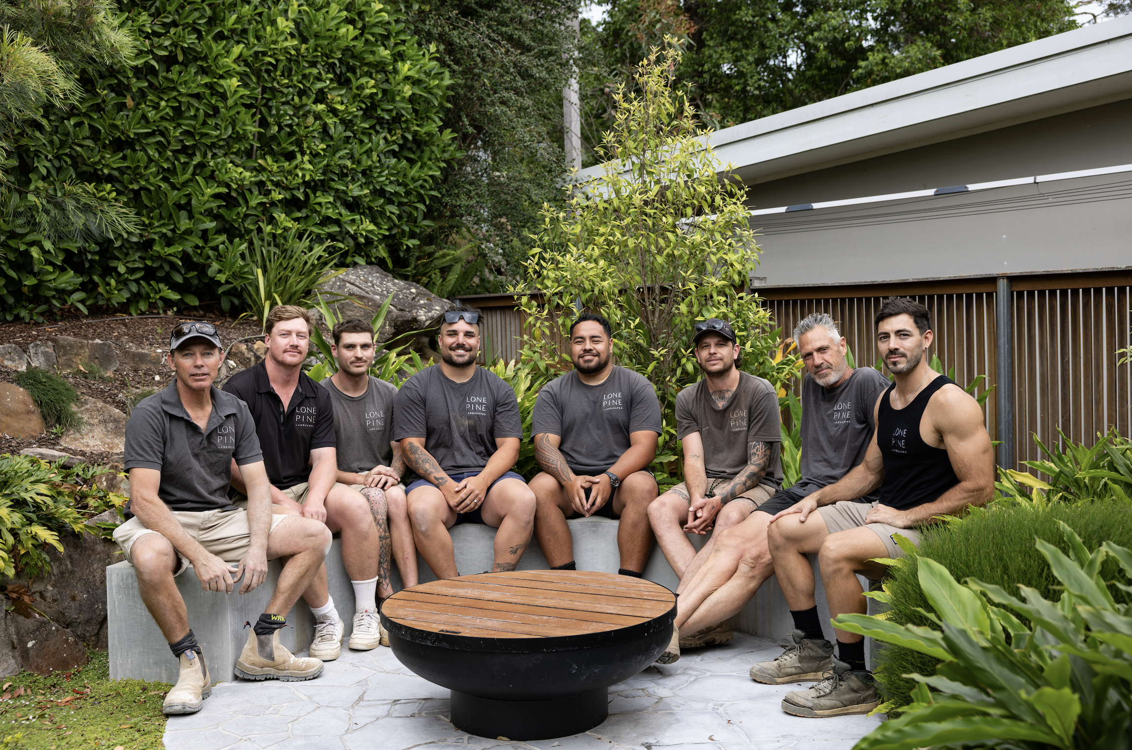 Group of eight men sitting outdoors on a concrete bench and around a firepit, surrounded by lush green plants and trees, with a modern building in the background.